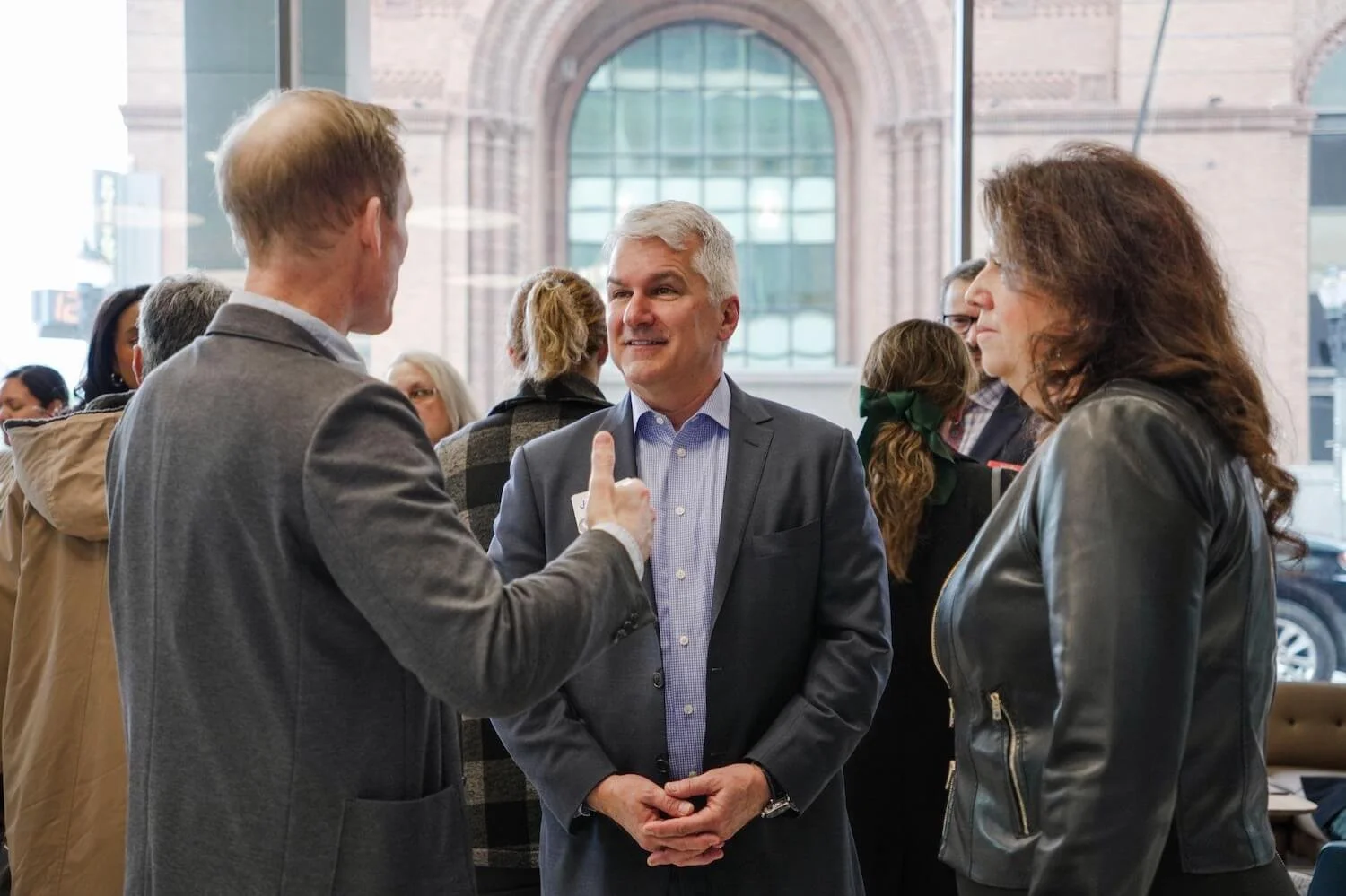 People engaged in conversation at an indoor event with a large arched window in the background.