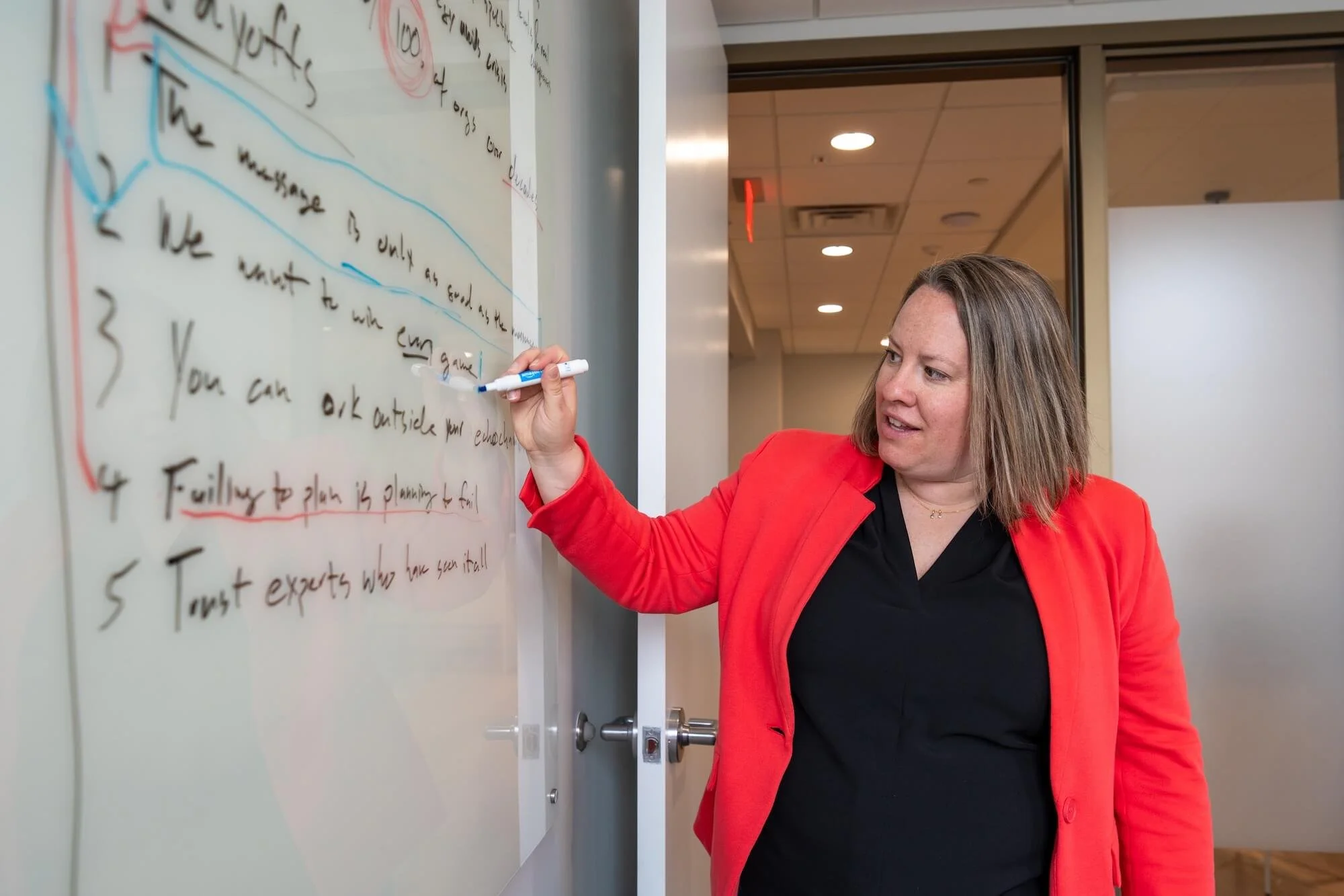 A woman with shoulder-length brown hair, wearing a black shirt and a red blazer, writing on a whiteboard with a blue marker in a conference room.