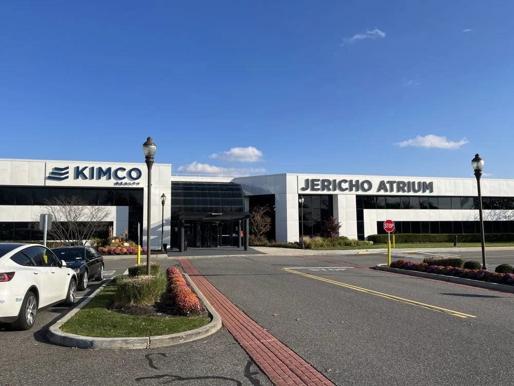 Exterior view of Jericho Atrium shopping center with Kimco signage, parking lot, and street lamps on a clear day