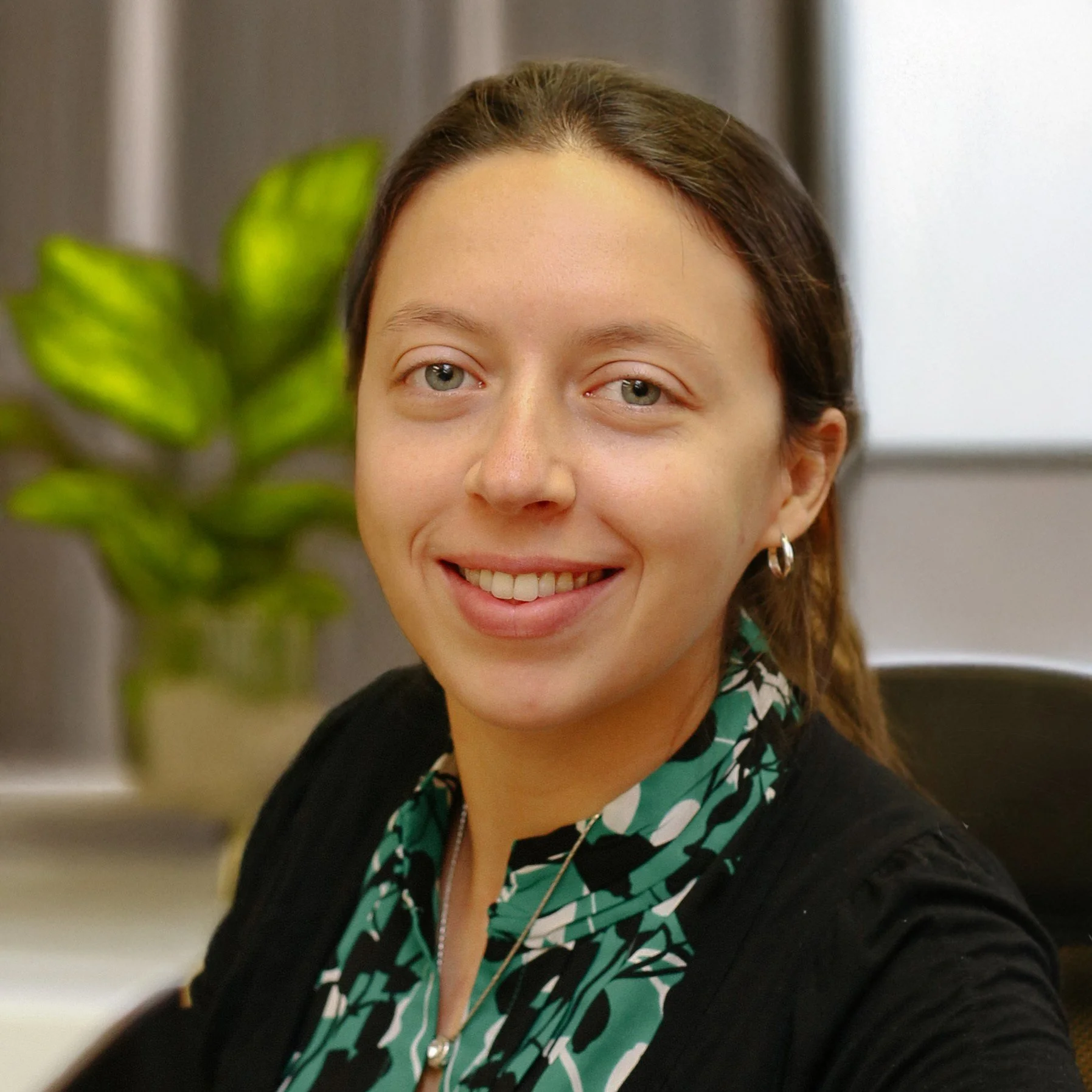 A woman with brown hair and green eyes smiling in an office setting, with a potted plant in the background.