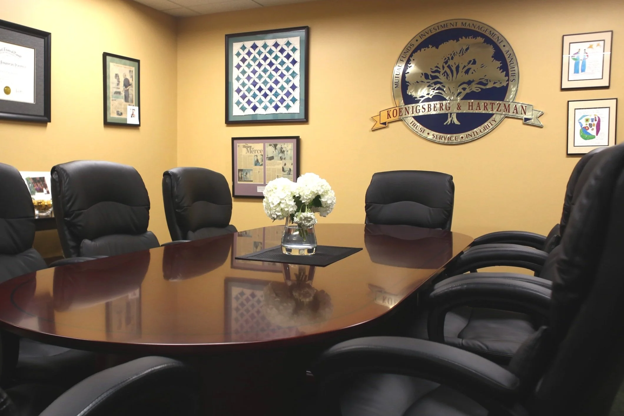 Conference room with a polished wooden table, six black leather chairs, a vase of white flowers, and framed artwork on yellow-painted walls. A large circular logo with a tree and the text 'Koenigsberg & Hartzman' is on the wall.
