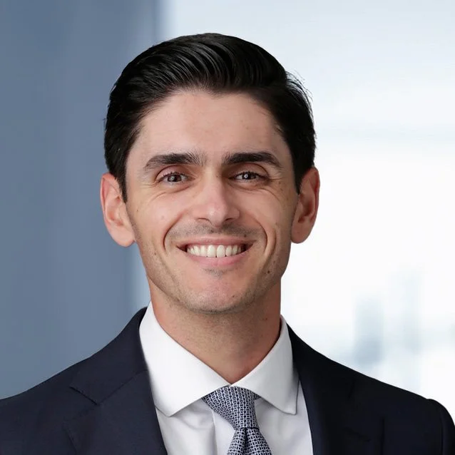 A young man with dark hair, wearing a navy business suit, white shirt, and patterned tie, smiling in an office setting with a blurred window background.