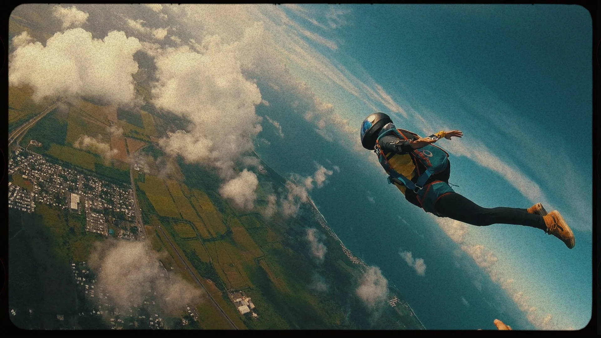 A skydiver in a helmet, harness, and yellow boots free-falling in the sky over a landscape with clouds, greenery, and a town below.