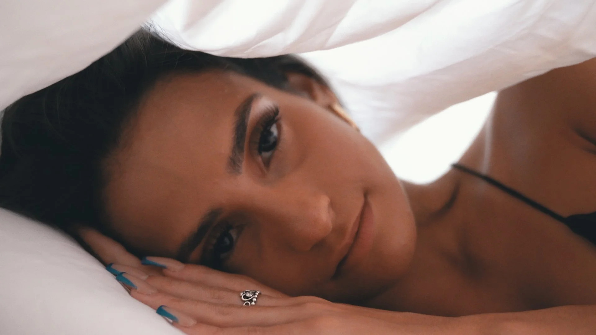 A woman lying on a bed, resting her head on her hands, wearing a black strap top, with makeup on her eyes and lips, and a ring on her finger.
