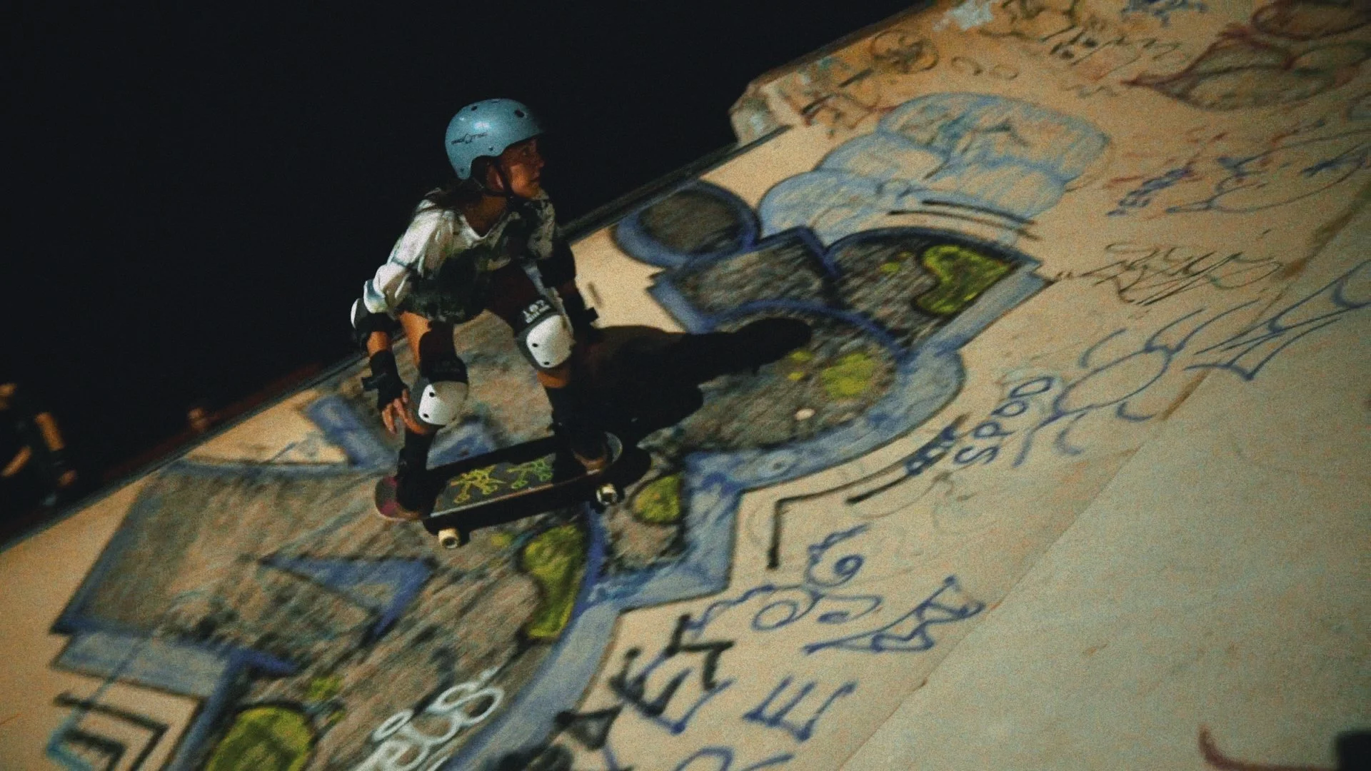 Skateboarder performing trick in front of graffiti art on an outdoor skate ramp at night.