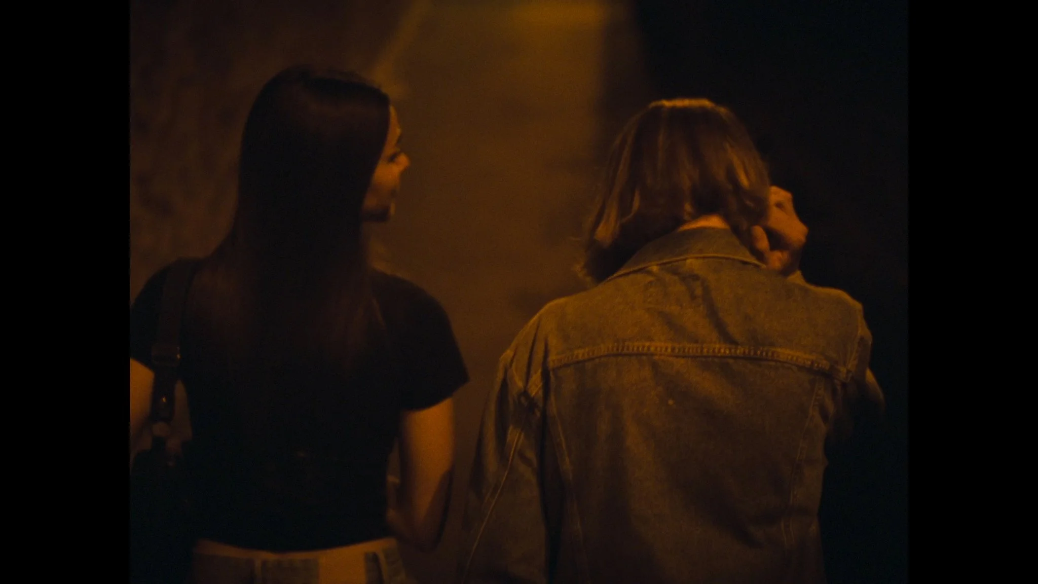 Two women standing close together in a dimly lit, possibly outdoor, setting. One woman has long dark hair and is wearing a black top, while the other woman has lighter, shoulder-length hair and is wearing a denim jacket. They are looking at each othe
