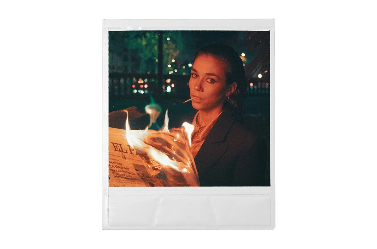 A woman with dark hair and a serious expression is smoking a cigarette, reading a newspaper, and holding a flaming object that looks like a magazine at night with blurred city lights in the background.