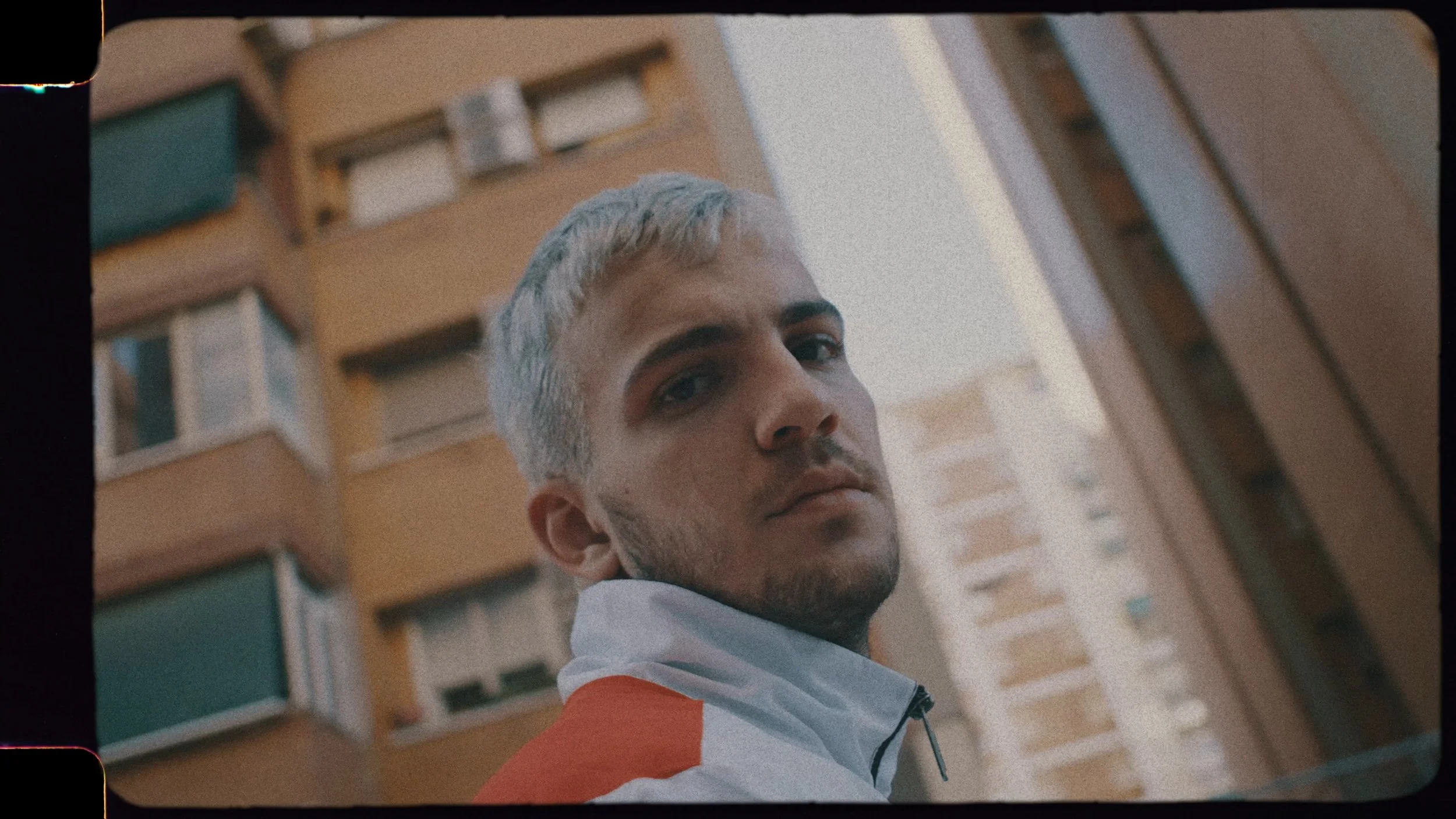 A young man with light gray hair and a beard looking at the camera, standing outdoors in front of a high-rise apartment building.