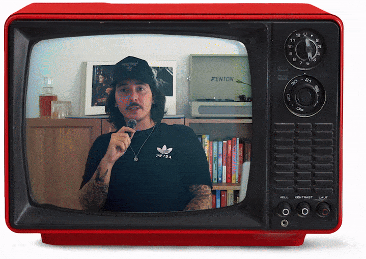 A man with dark long hair, mustache, and tattoos on his arms is speaking into a microphone, reflected in a vintage red television screen. Behind him are books on a shelf, a framed picture, and a Fenton amplifier.
