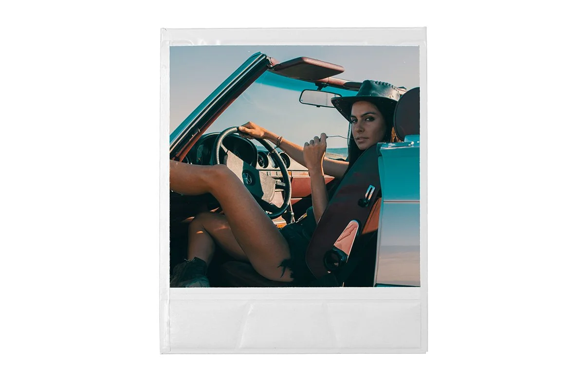 Woman wearing a hat sitting in a vintage convertible car, holding the steering wheel and looking at the camera, with a clear sky background.