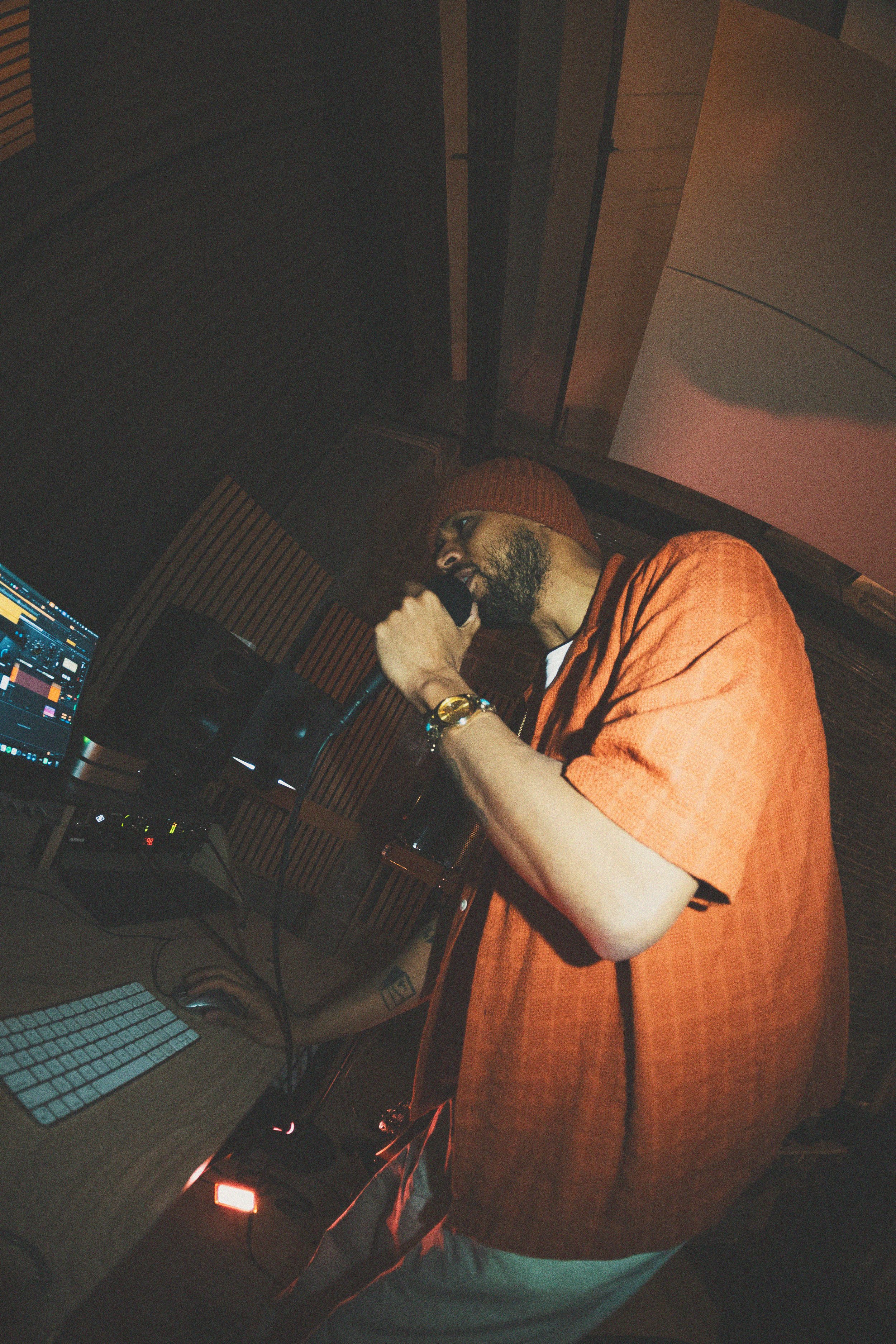 A man wearing an orange beanie and matching orange shirt, with a beard, holding a microphone in a recording studio.