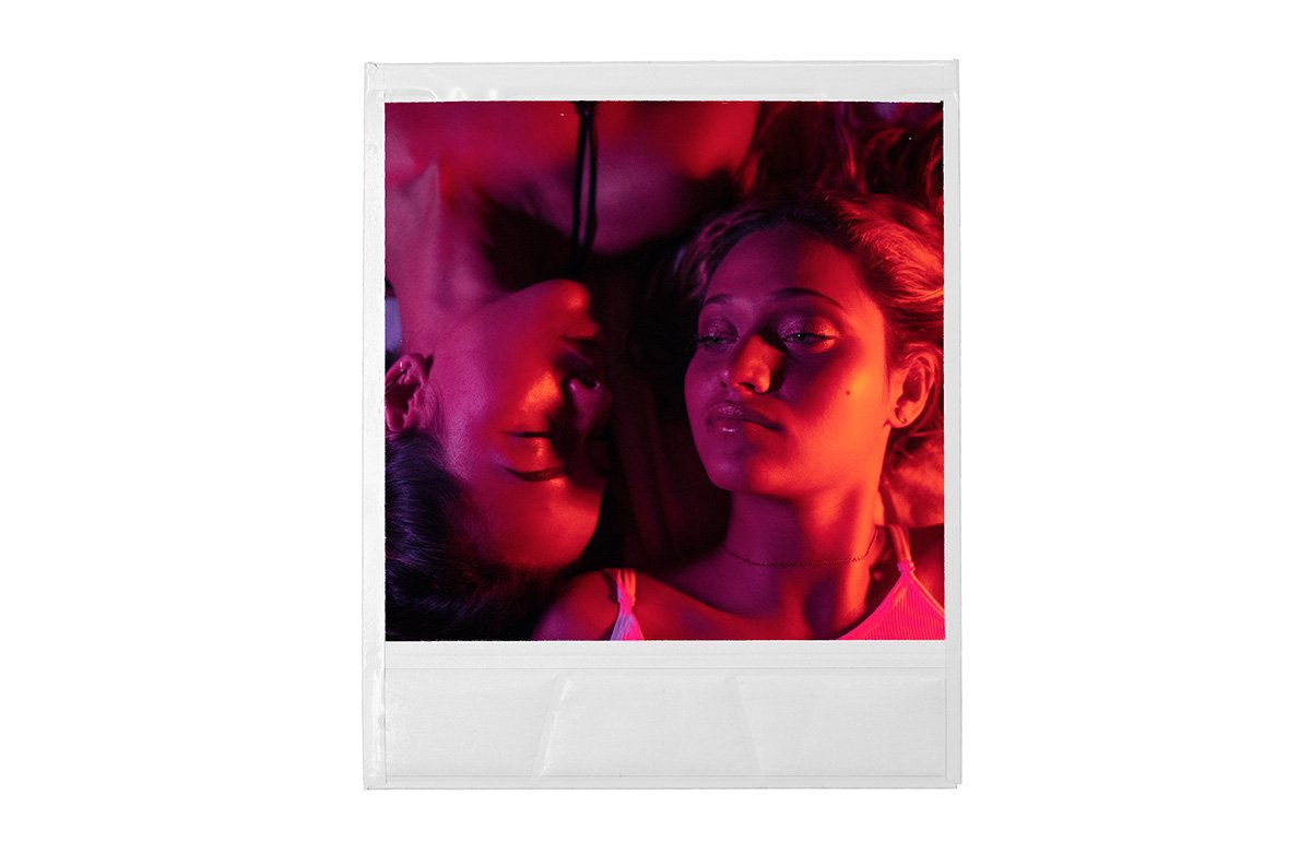 Polaroid photo of two women lying down, illuminated by pink and purple lighting, one with eyes closed and the other looking to the side.