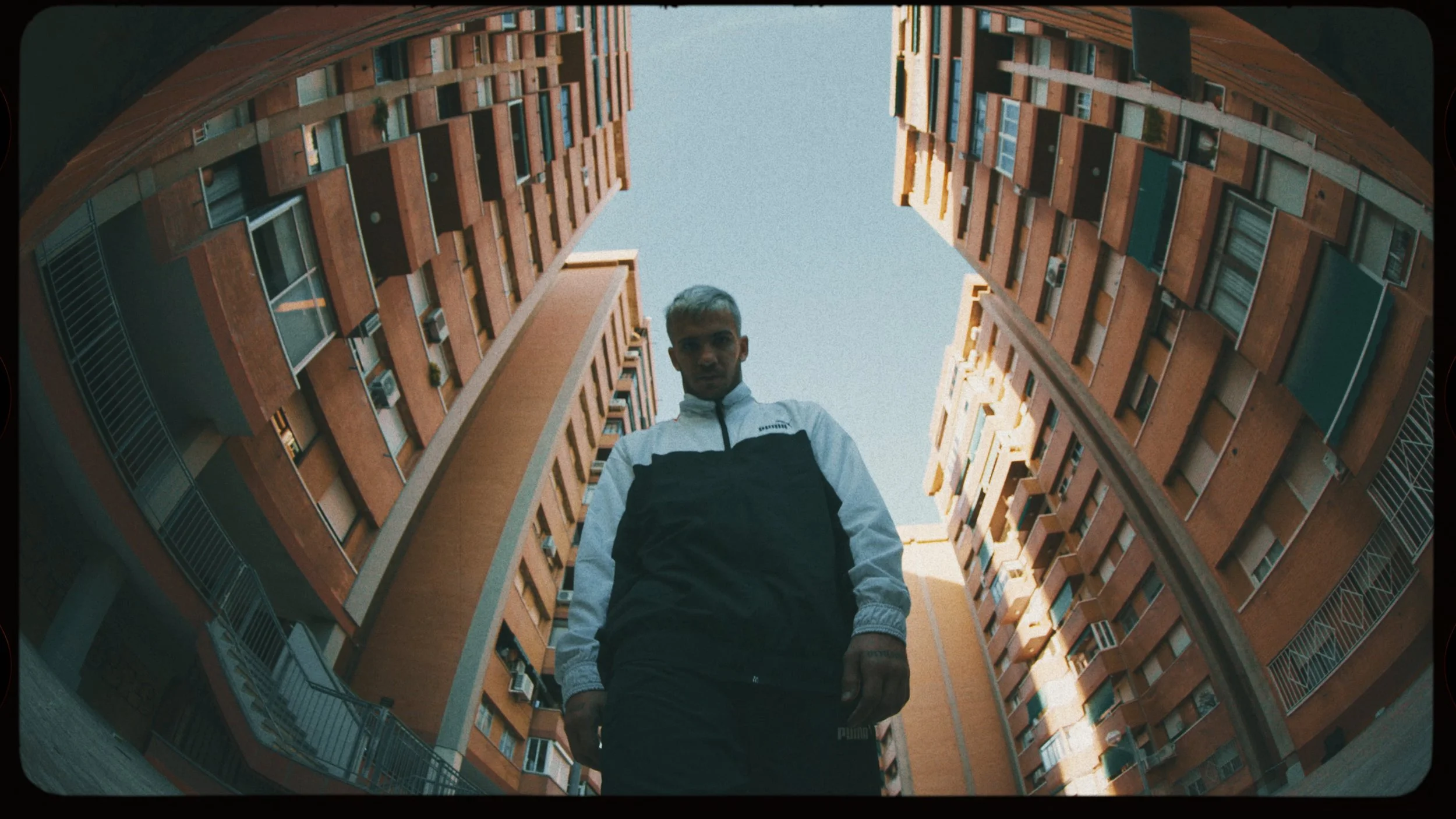 A young man with bleached hair wearing a black and white Puma jacket stands in the courtyard of tall orange brick apartment buildings, looking down at the camera from a low angle.
