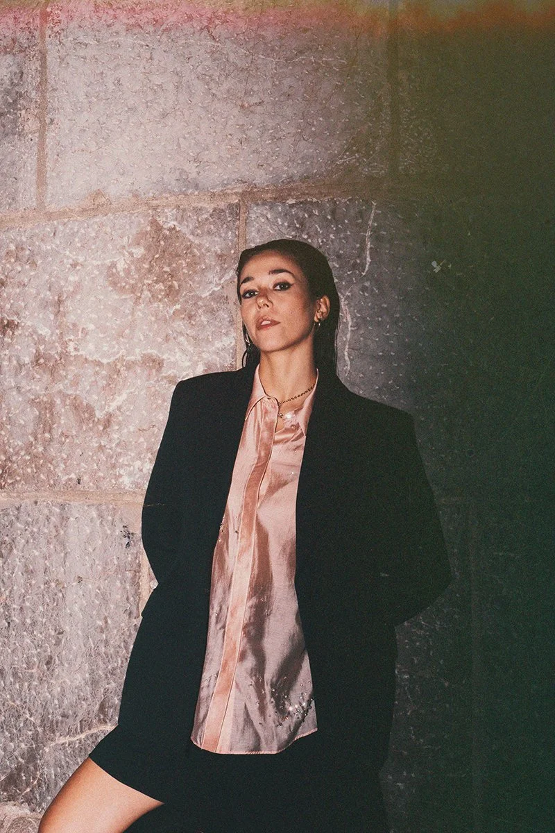 A woman with dark hair wearing a pink silk blouse, a black blazer, and black shorts standing against a stone wall.