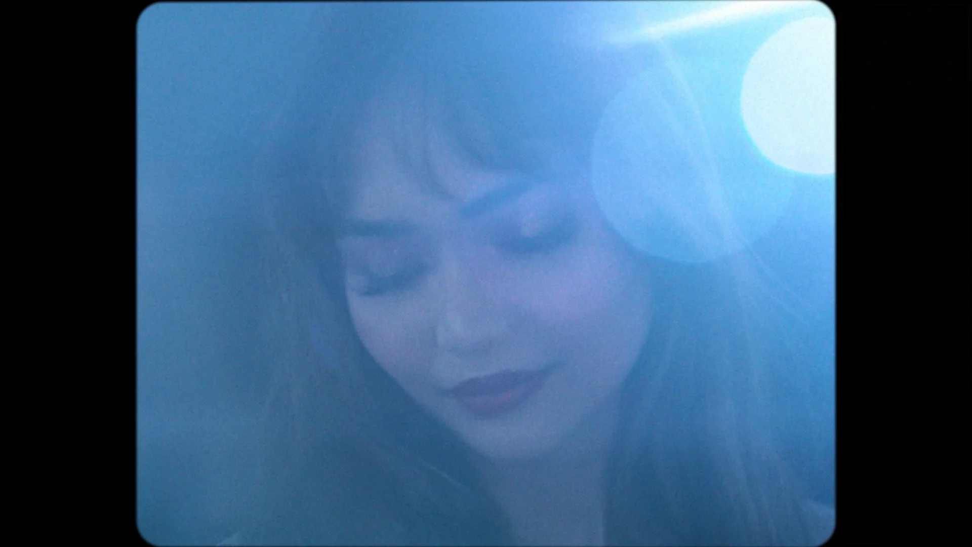 A woman with dark hair and makeup, eyes closed, appears to be looking downward, with a blue filter overlay and lens flare effect in the background.