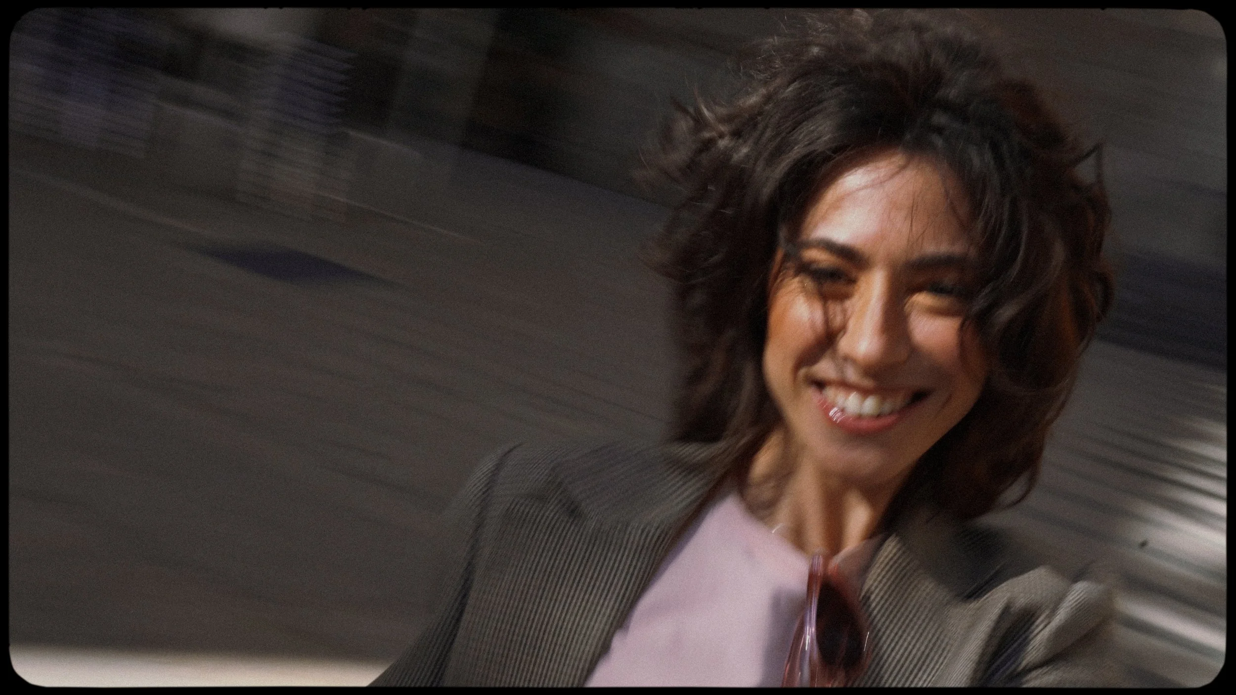 Close-up of a woman with wavy dark hair smiling and winking, wearing a gray blazer and pink top, with blurred background.