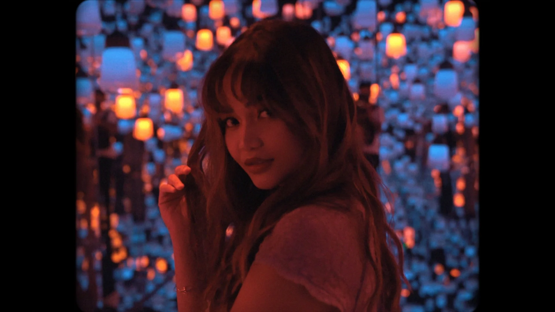A woman with long hair, wearing a pink top, is illuminated by neon lighting. The background is filled with numerous small lights in orange and blue hues, creating a vibrant, colorful scene.