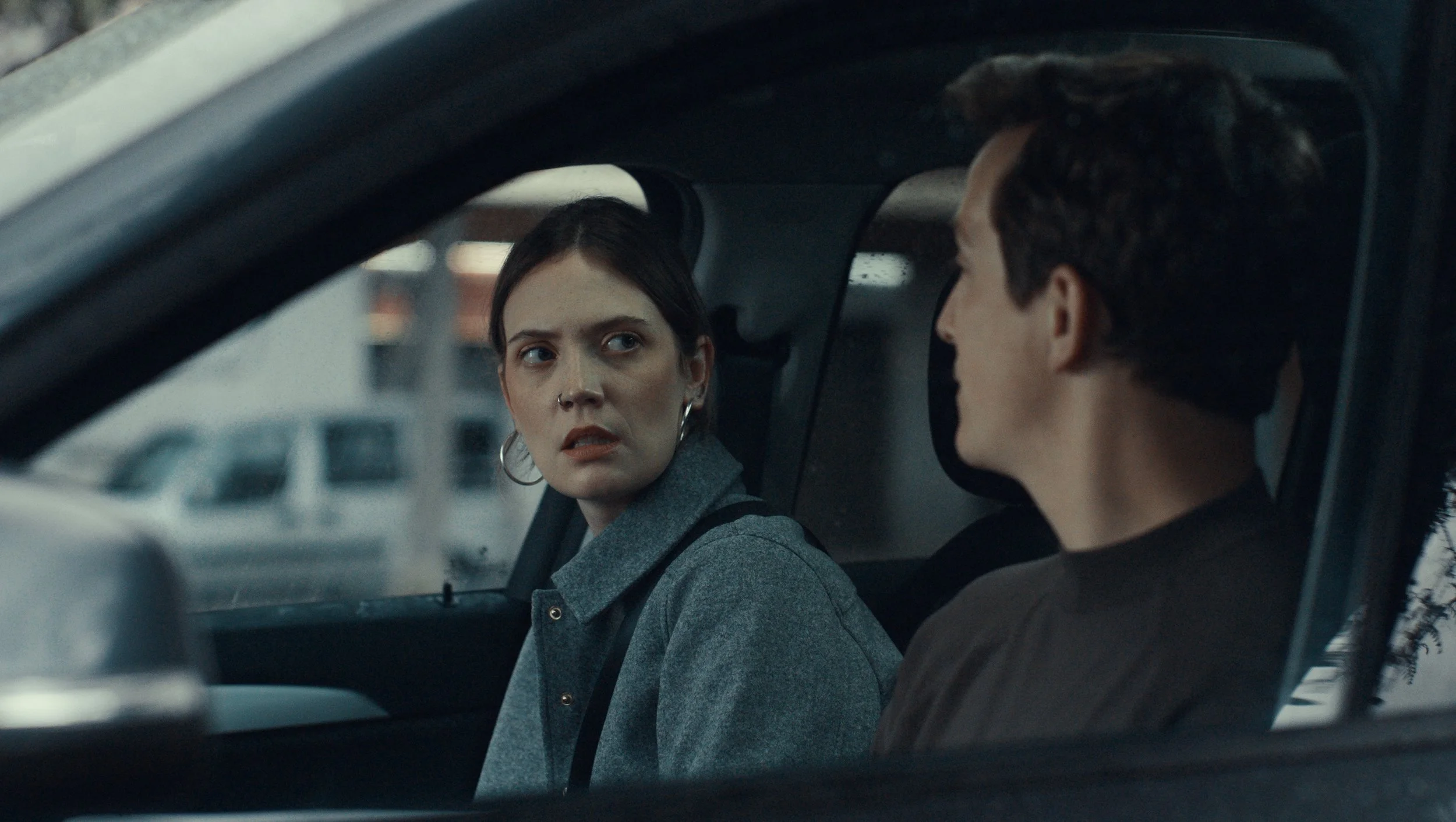 A woman with dark hair talking to a man with dark hair inside a vehicle. The woman has a concerned or confused expression.