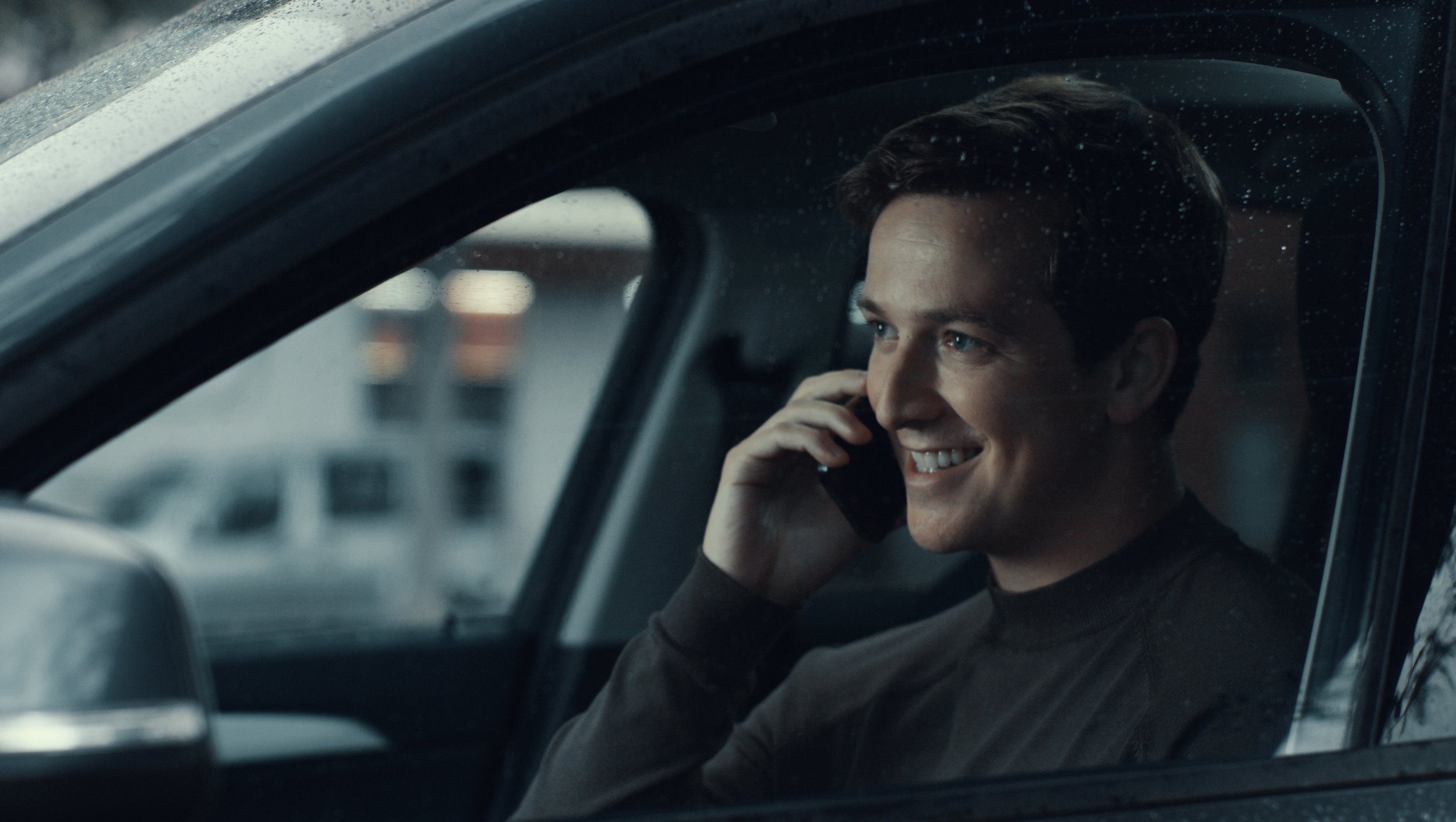 A young man with dark hair smiling while talking on a mobile phone inside a vehicle during rainy weather