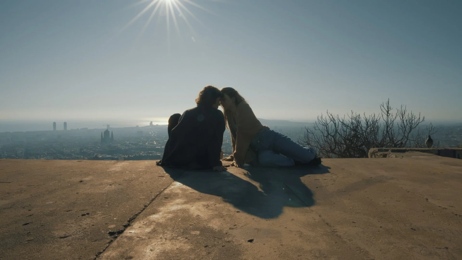 Two people sitting on a ledge with a city skyline in the background, sharing a moment and touching foreheads in front of a bright sun.