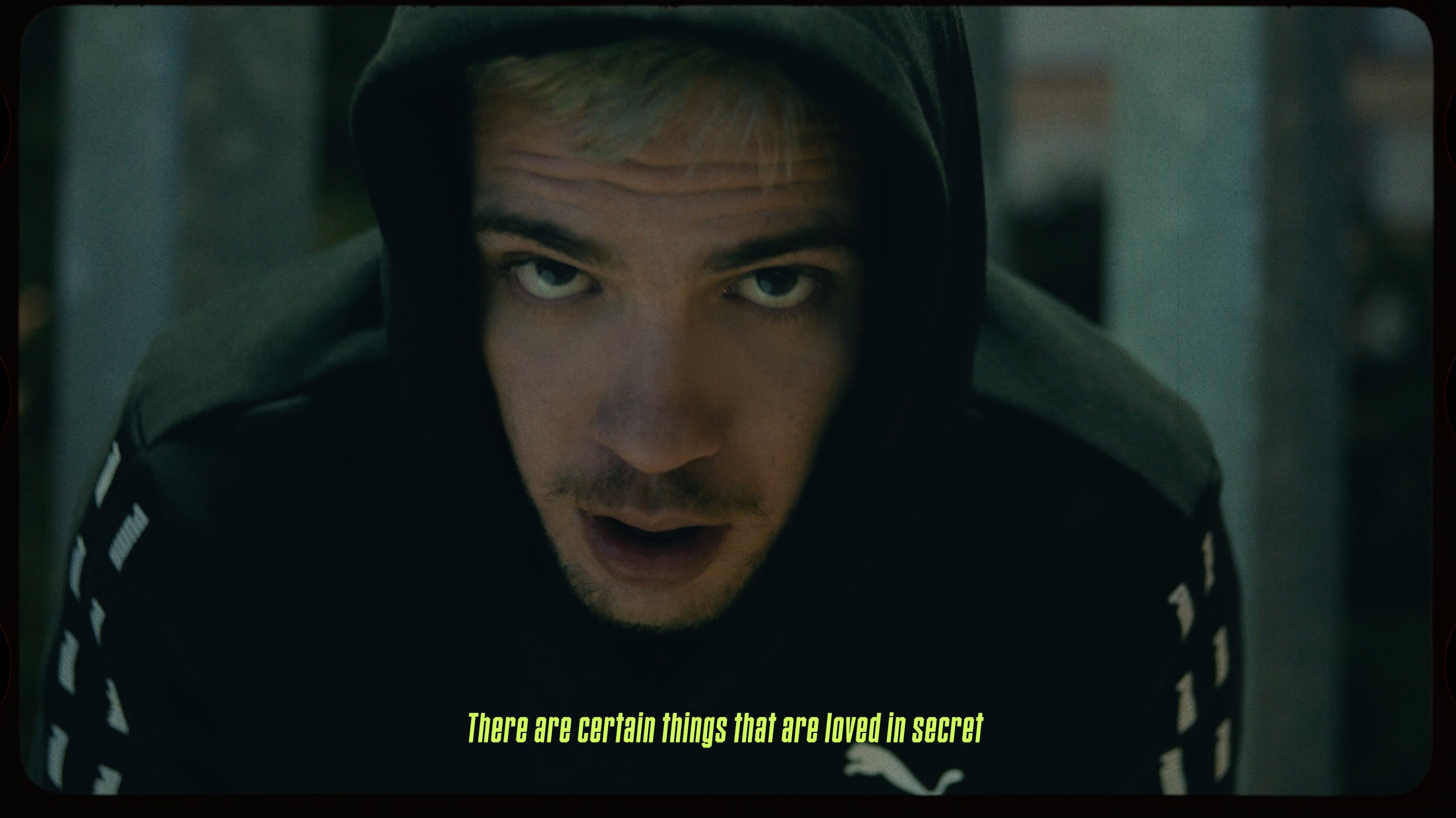 Close-up of a man with light hair, blue eyes, and slight facial hair, wearing a black hoodie with gray accents, staring intensely at the camera. There are yellow subtitles at the bottom that read, 'There are certain things that are loved in secret.'