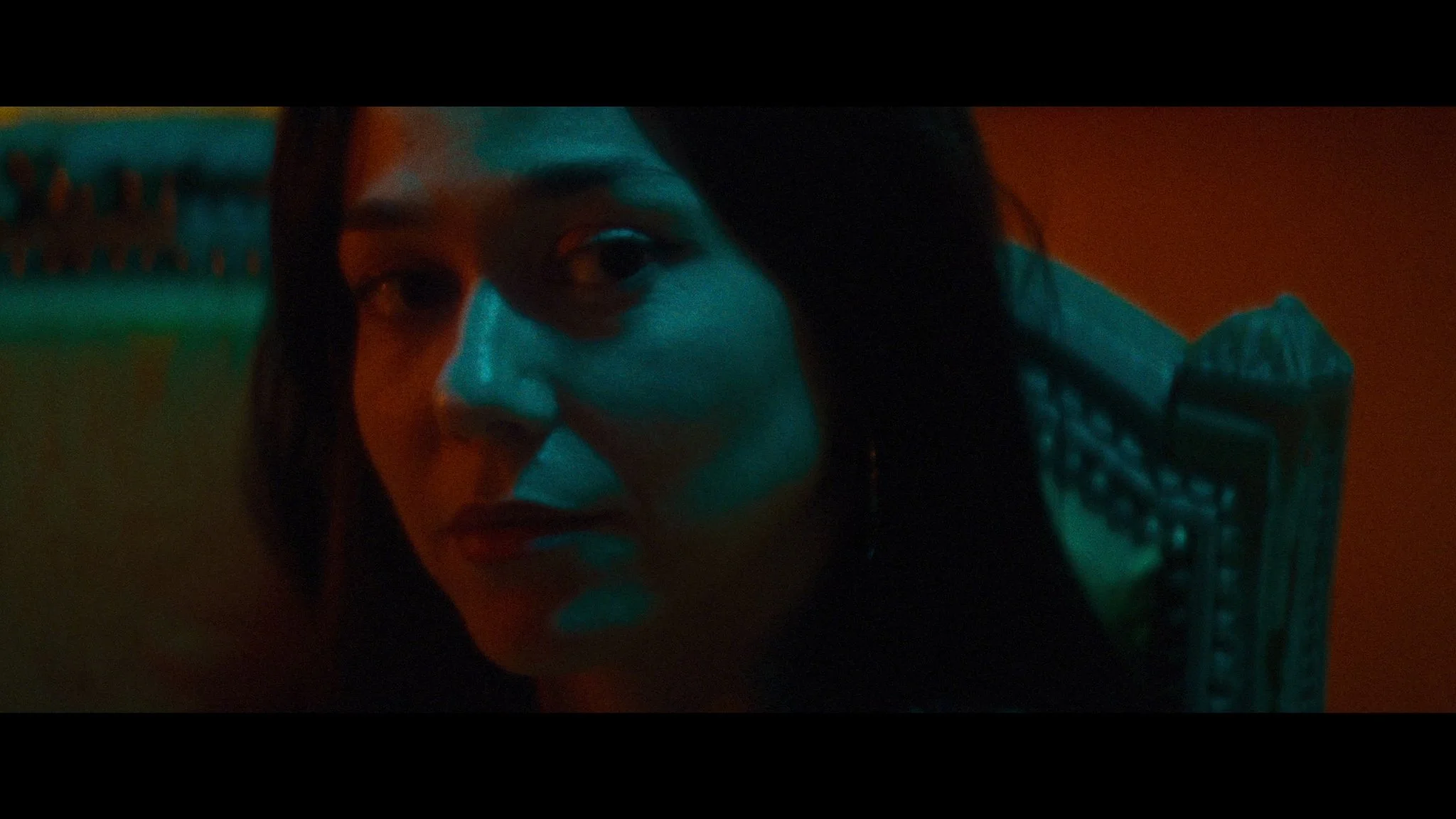 Close-up of a woman with dark hair sitting on a wooden chair in dim, colorful lighting, looking over her shoulder.