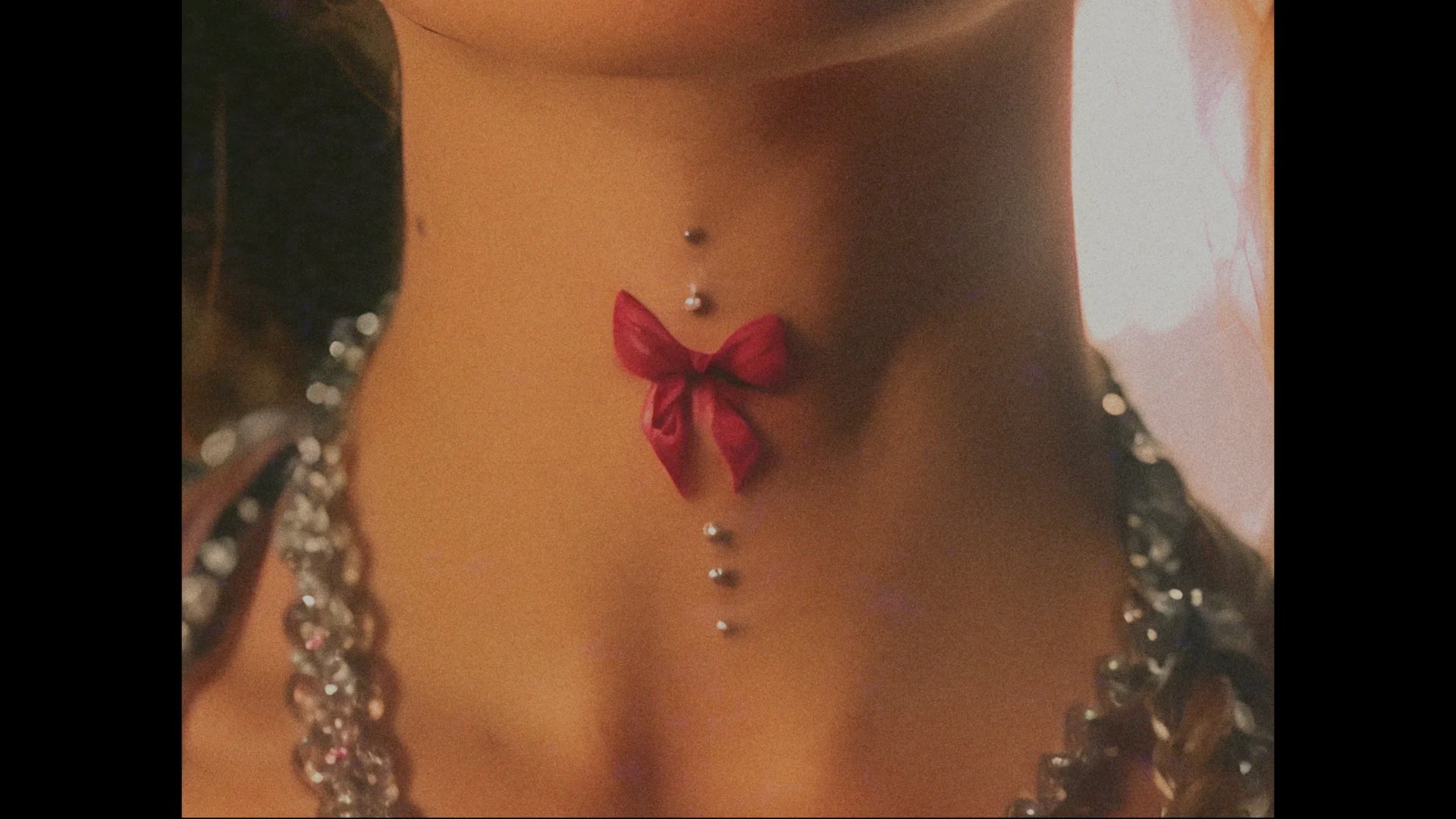 Close-up of a woman's exposed midriff with a red bow and belly button piercing, surrounded by sparkly jewelry.