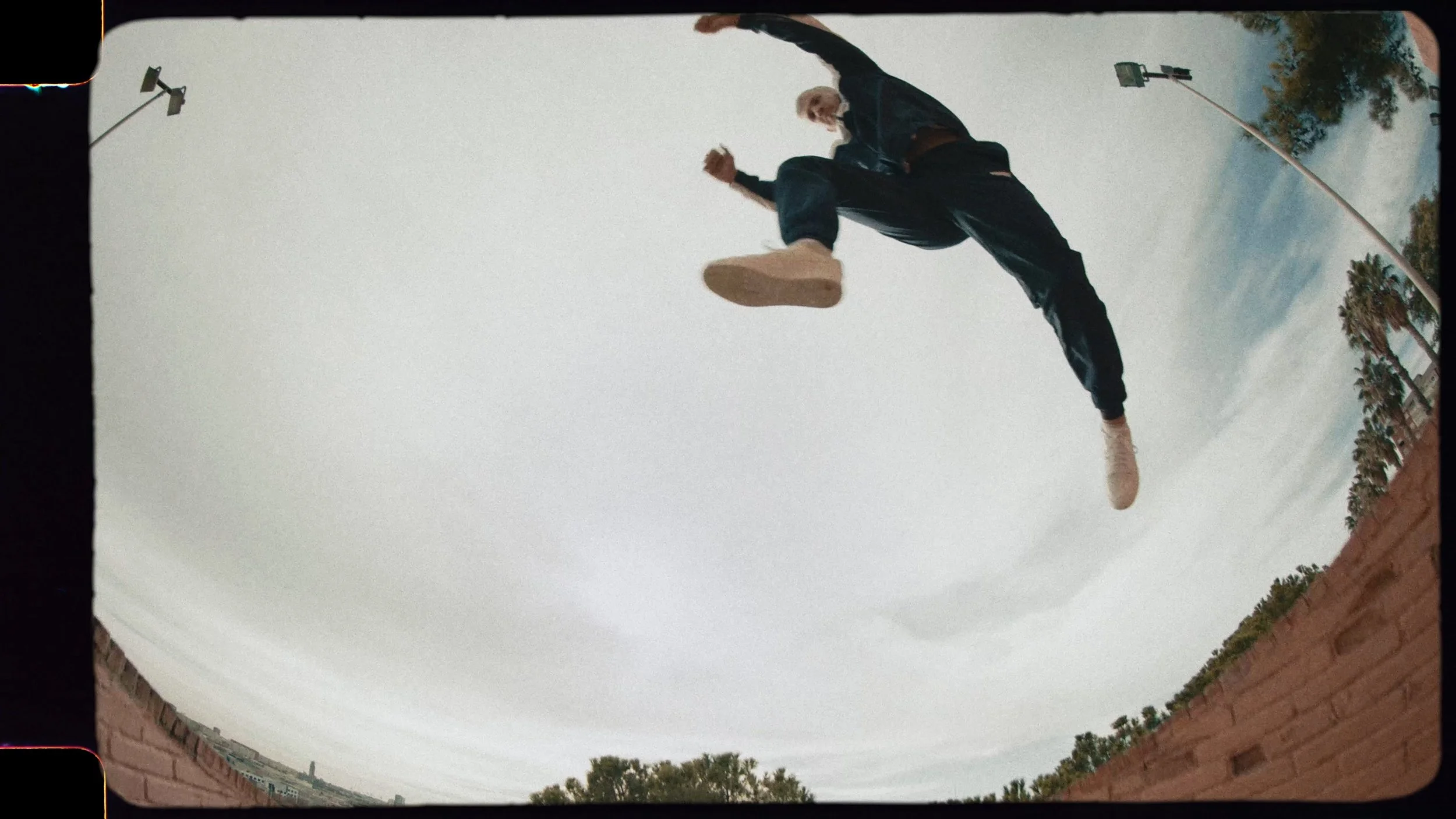Person jumping in the air outdoors, captured in a fisheye lens perspective, with trees, cloudy sky, and a brick wall in the background.