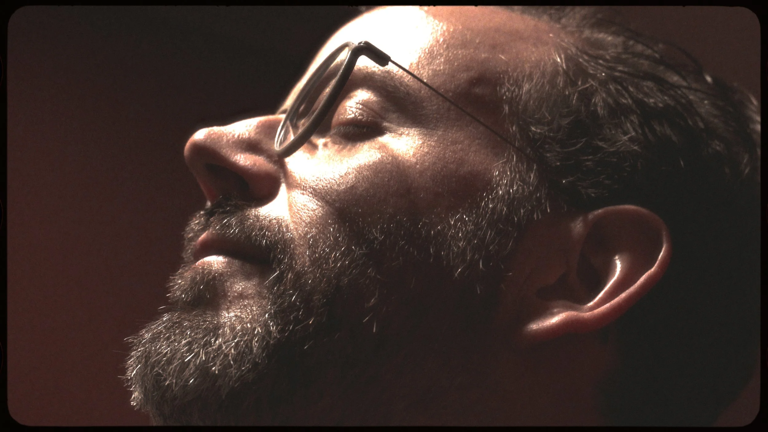 Close-up side profile of a man with sunglasses and a thick beard, with soft lighting highlighting his features.
