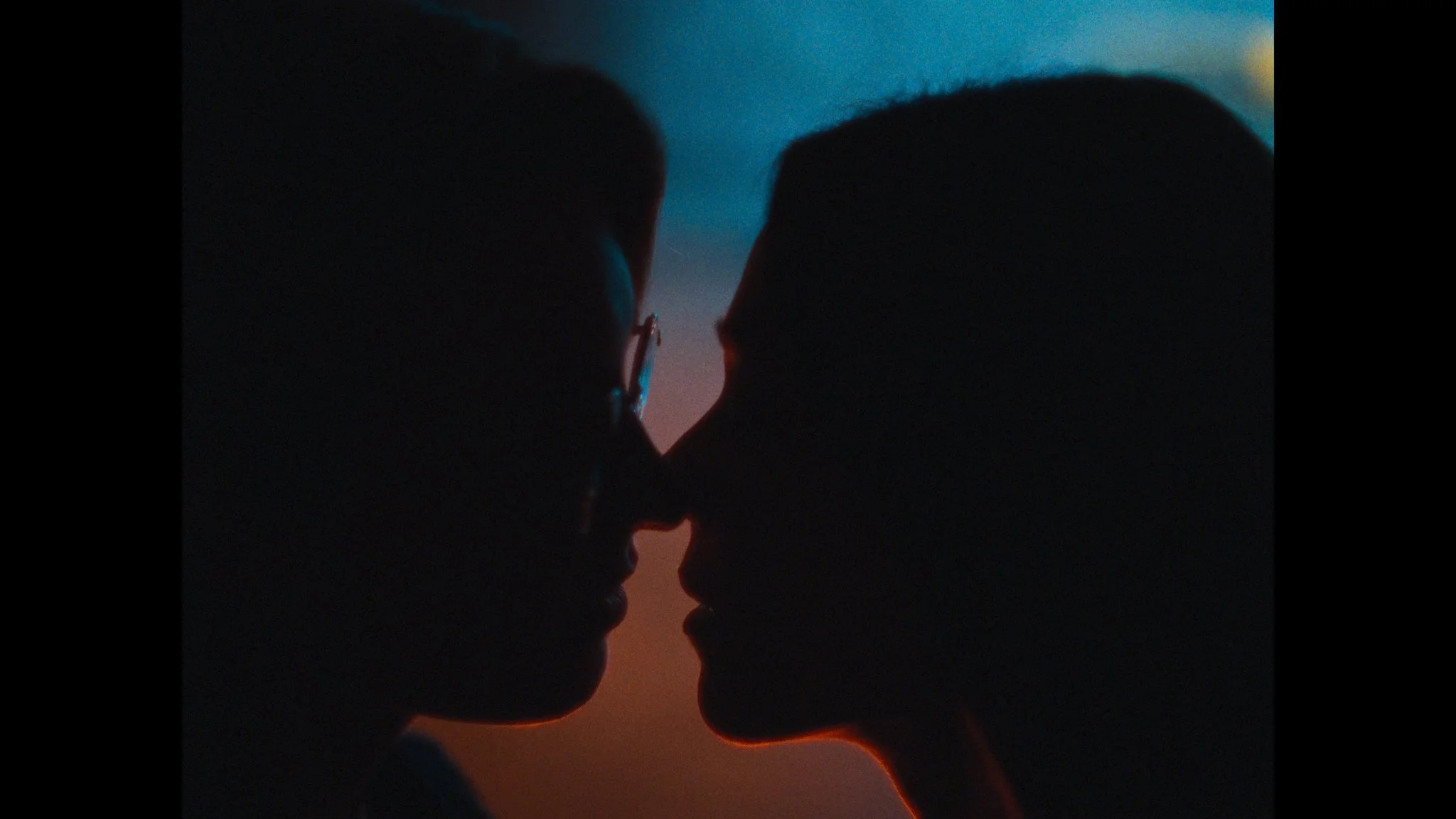 Silhouette of a couple face-to-face with their noses nearly touching, with a blurred background of sunset or twilight colors.