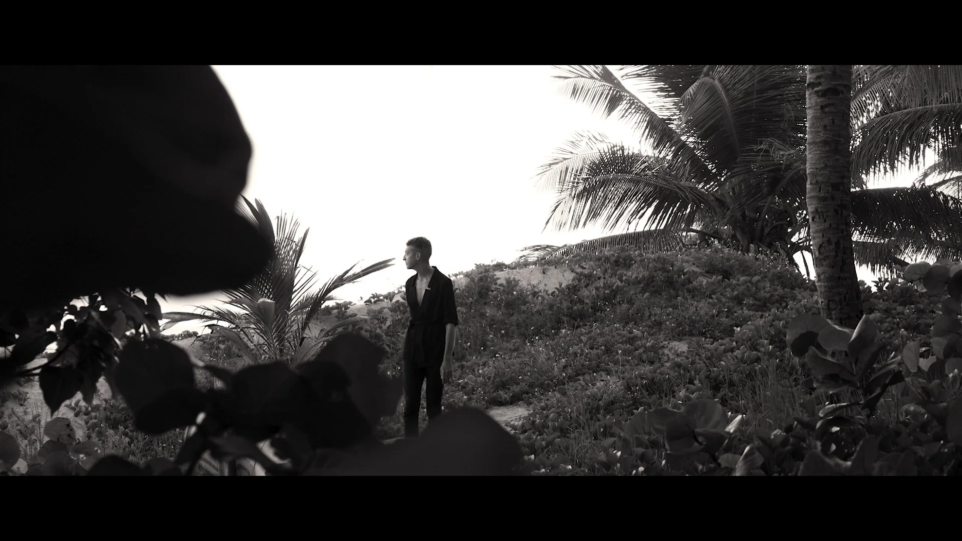 A woman standing in a tropical setting with lush vegetation and palm trees, seen through a frame of dark foliage in the foreground, in black and white.
