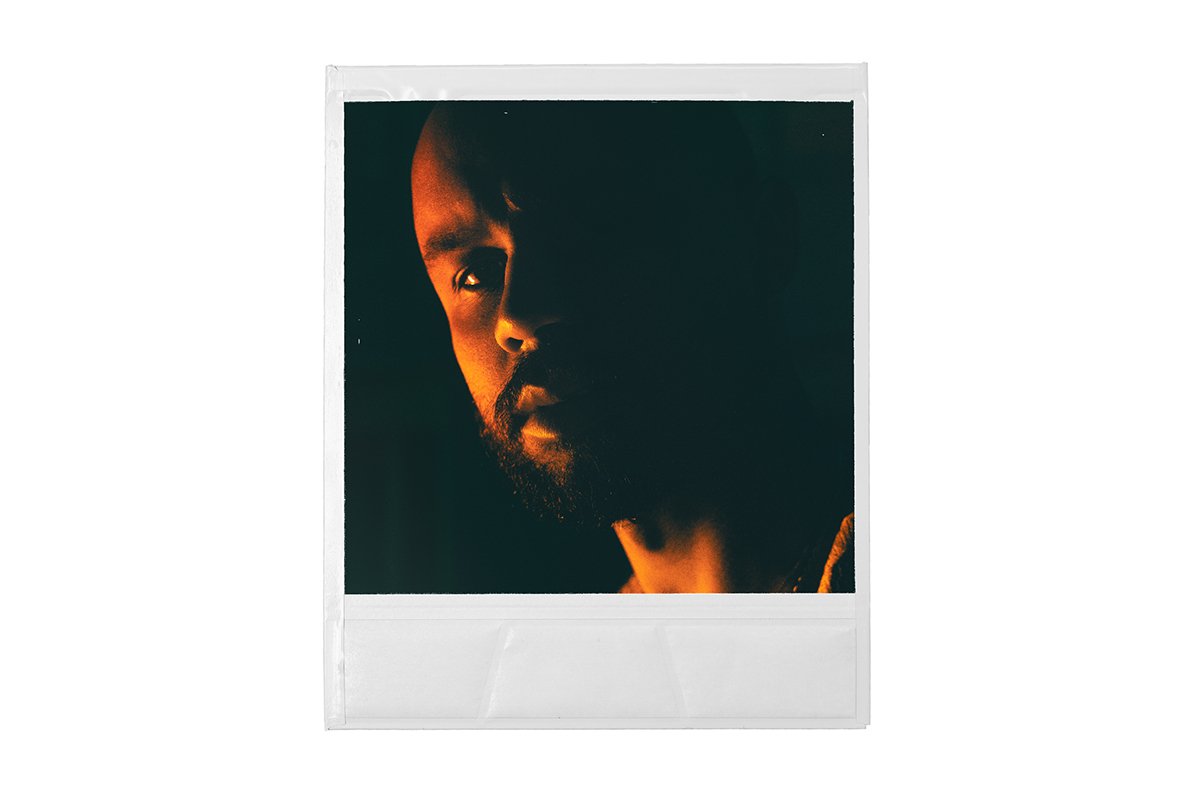 Close-up portrait of a man with dark hair and a beard, partially illuminated by warm lighting, against a dark background, resembling a vintage instant photo style.