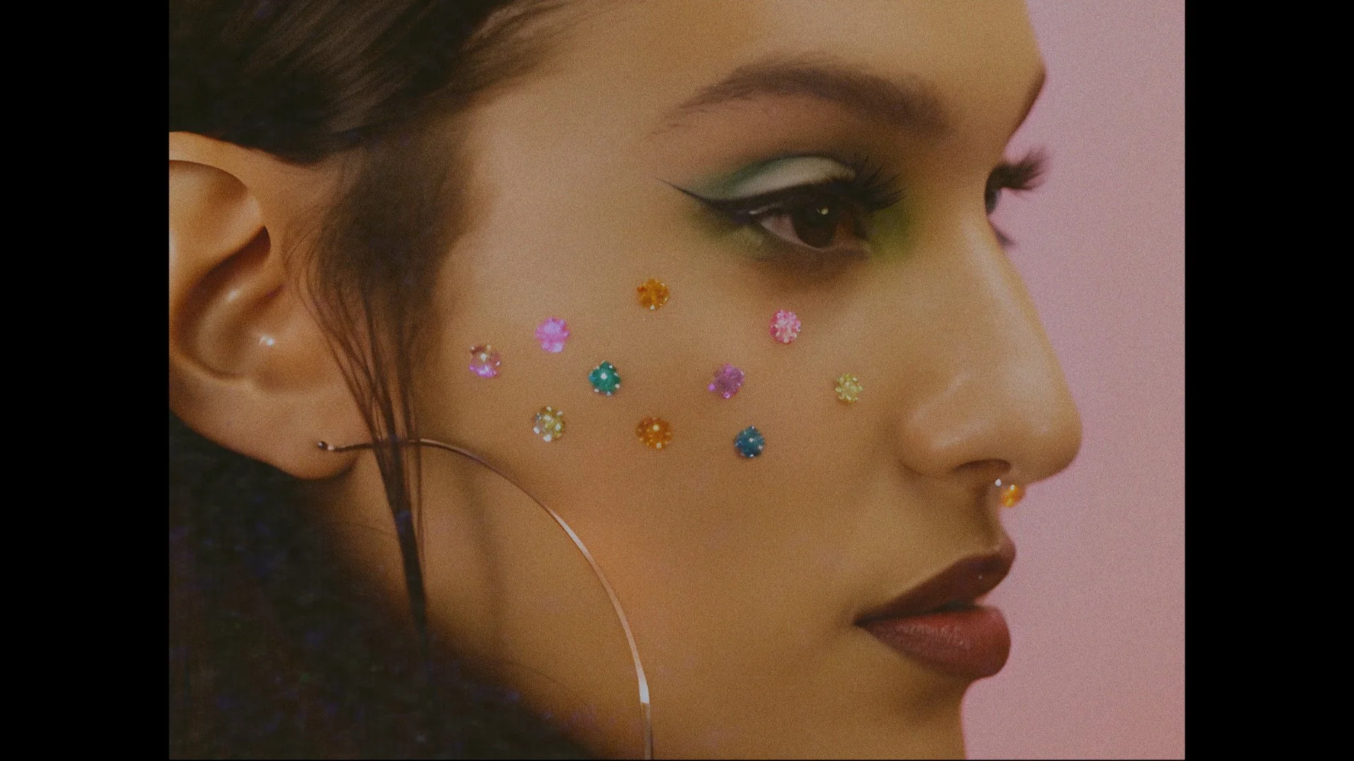 Close-up of a woman's face with colorful flower-shaped rhinestones on her cheek, dark hoop earring, and makeup including green eyeshadow and black winged eyeliner.