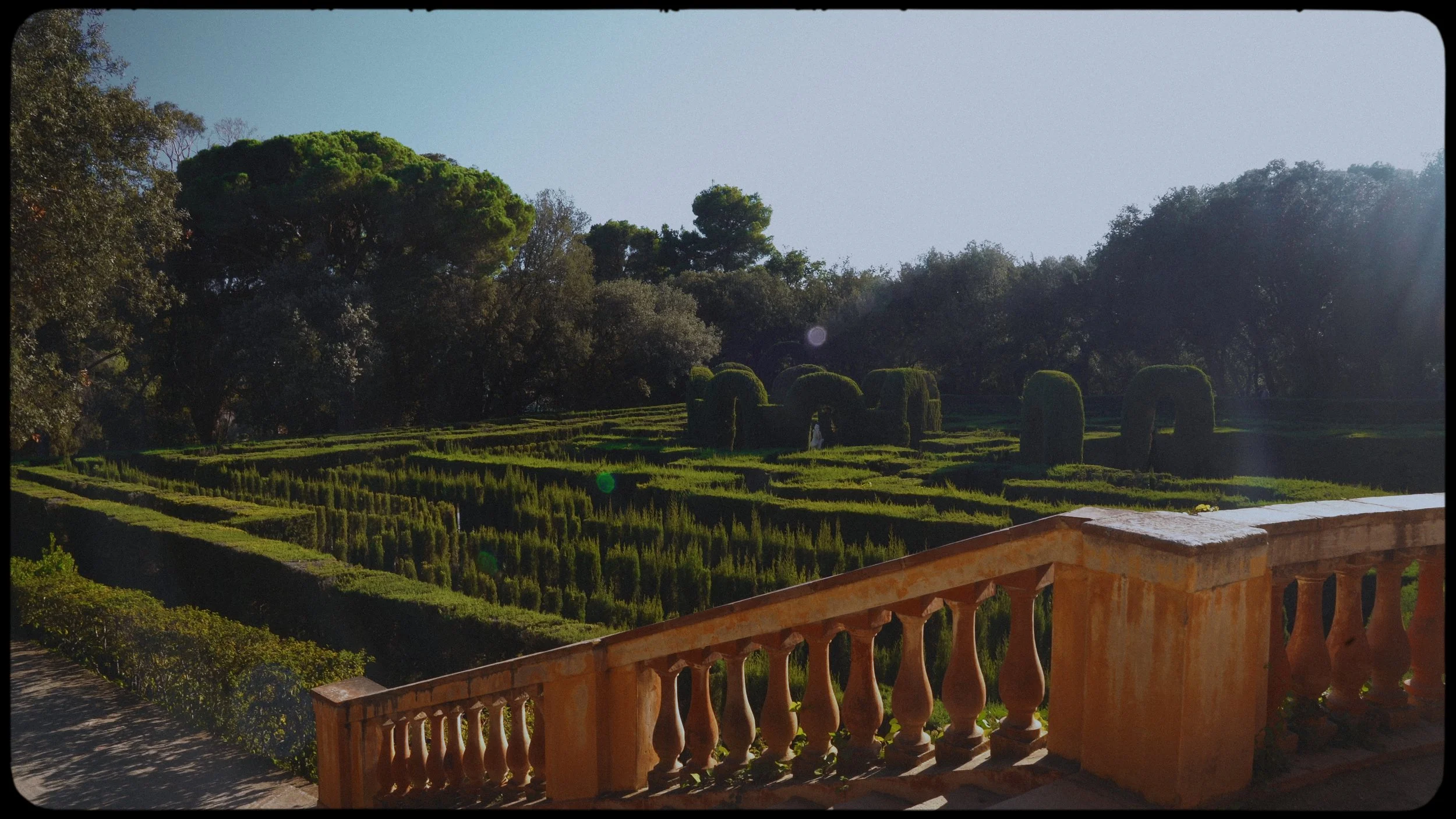 Photograph of a landscaped garden with trimmed hedges, topiary shaped in arches, and trees in the background, taken from a terrace with a brick railing, during daylight.