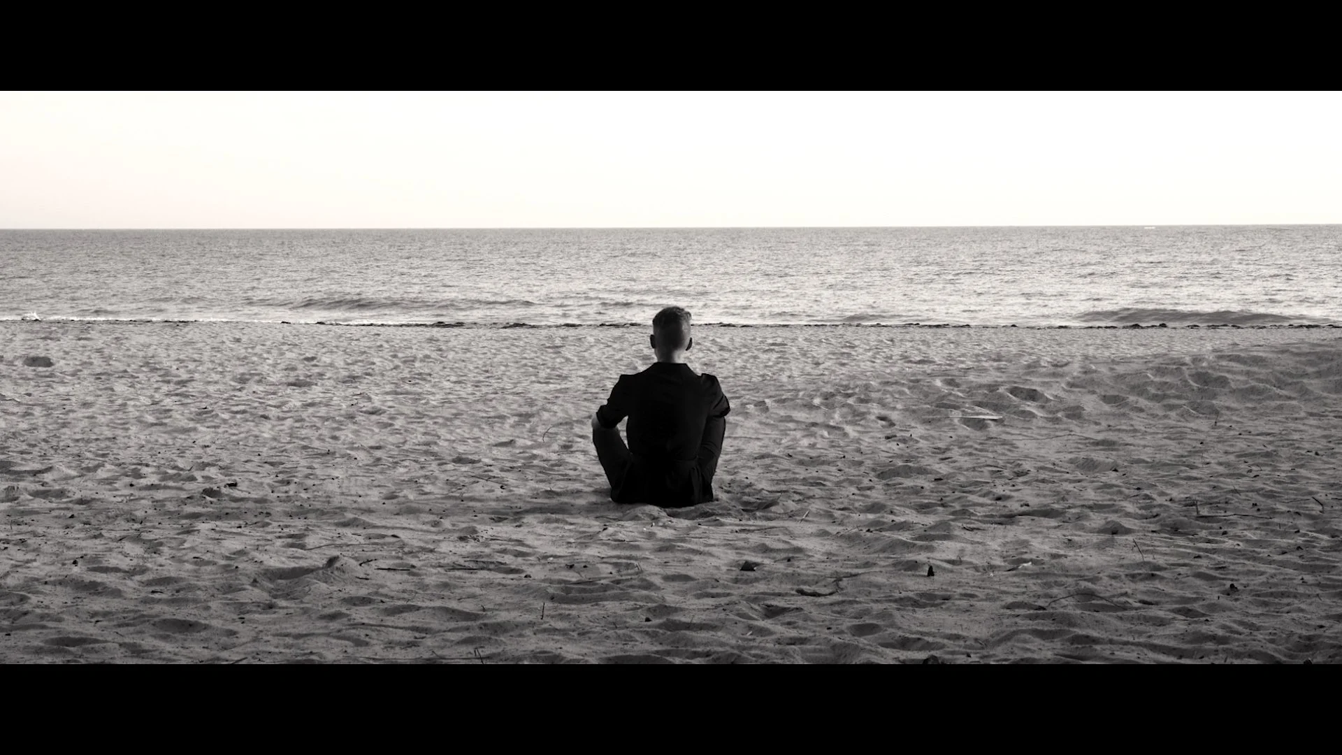 A black and white photo of a person sitting on a sandy beach facing the sea, with a calm ocean in the background, practicing meditation or contemplating.