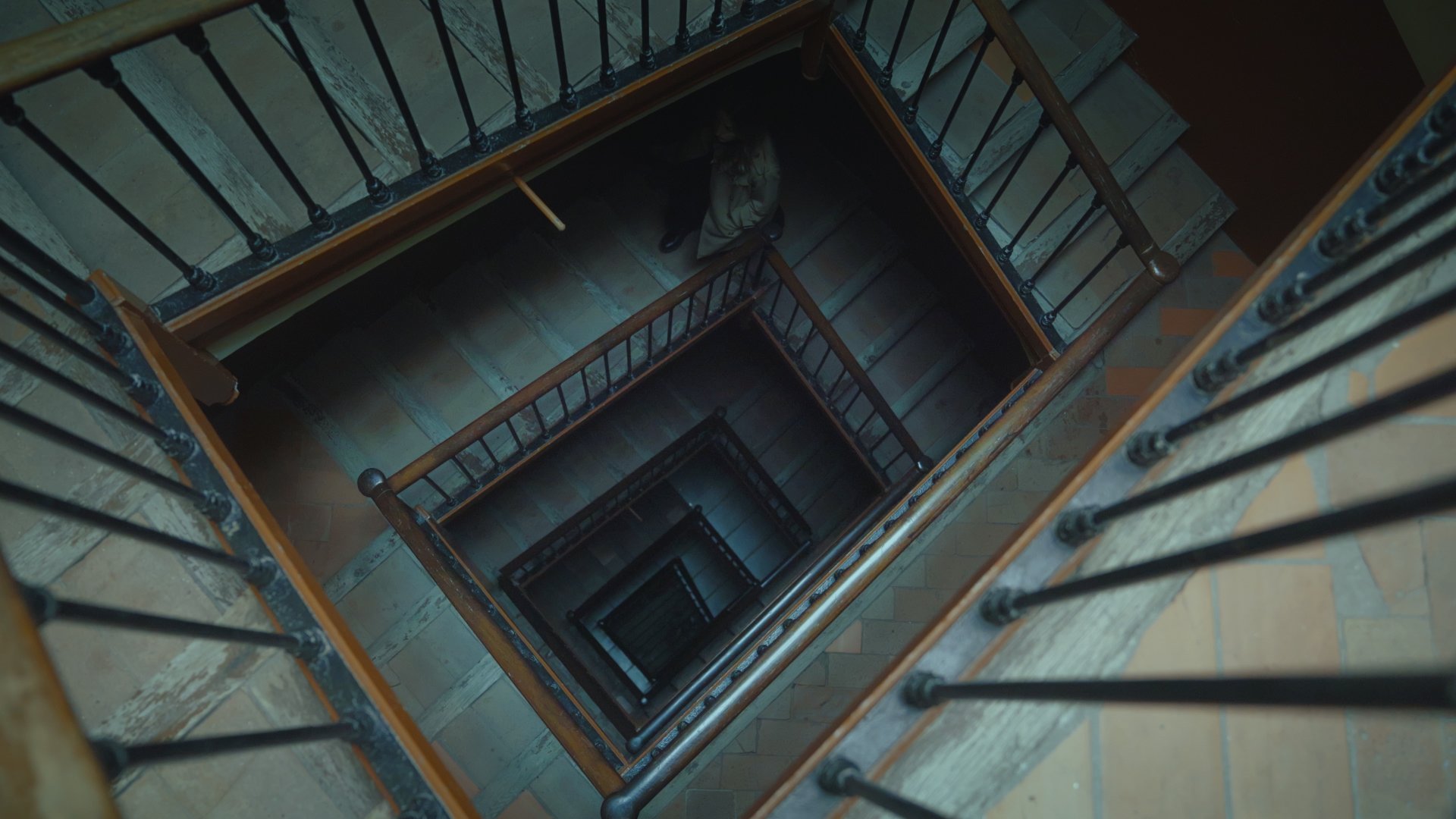 View looking down a multi-story staircase with wooden railings and black iron balusters, showing the spiral pattern of the stairs descending into darkness.