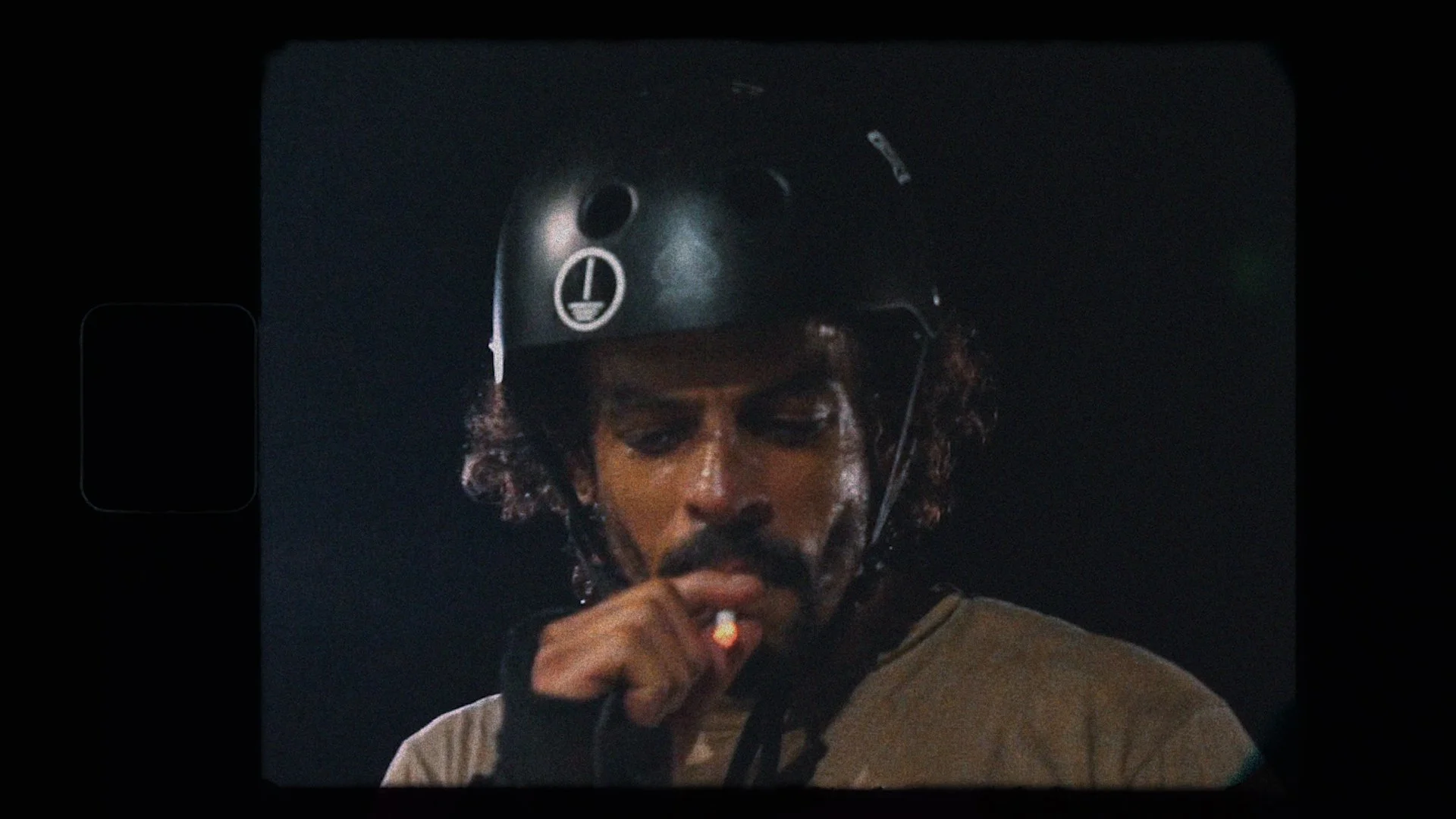 A man with dark curly hair and a mustache, wearing a black helmet, smokes a cigarette against a dark background.