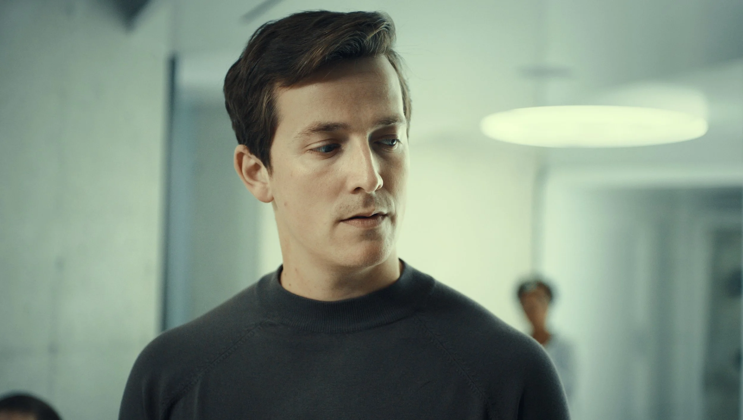 A young man with brown hair looking to his right, wearing a black crewneck shirt, in a modern indoor setting with a circular ceiling light and blurred background.