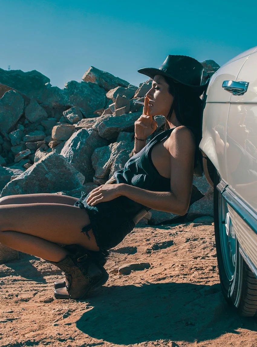 A woman sitting on the ground with her back against a white classic car, wearing a black hat, sleeveless top, and shorts, with rocky terrain and blue sky in the background.