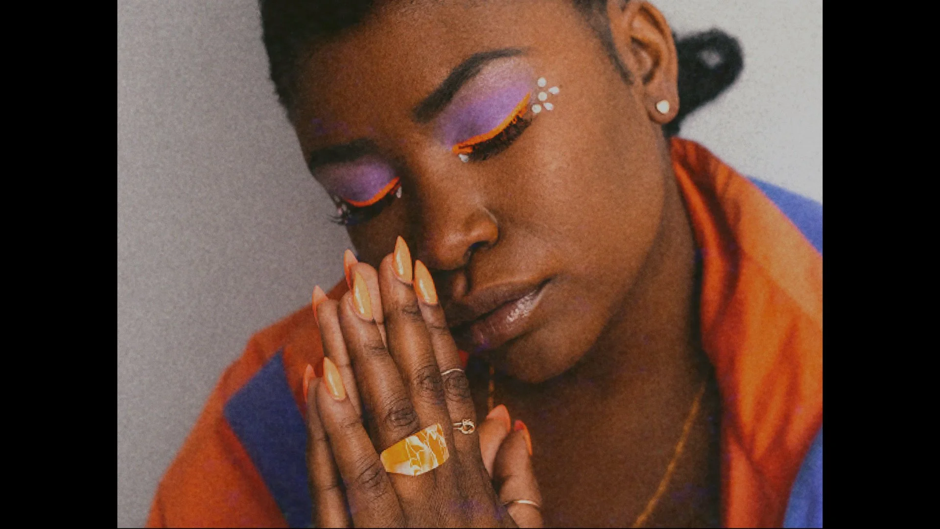 A woman with closed eyes, vibrant purple eyeshadow, orange eyeliner, and orange nail polish, holding her face with her right hand, wearing rings, earrings, and a ring on her left hand, with a gold chain necklace, dressed in an orange and blue garment