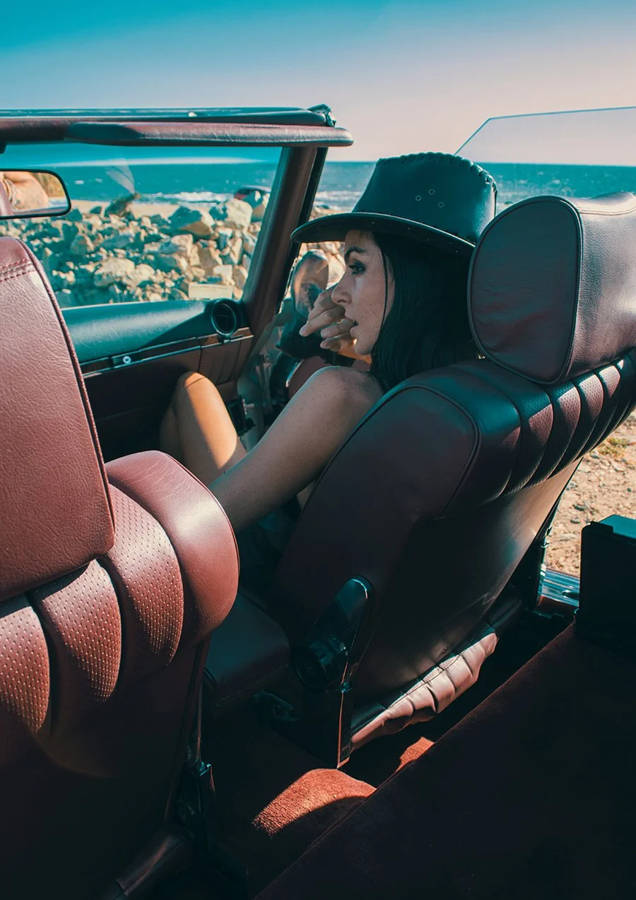 A woman sitting in a vintage convertible car with black leather seats, wearing a black hat, near a rocky beach with the ocean and a clear sky in the background.
