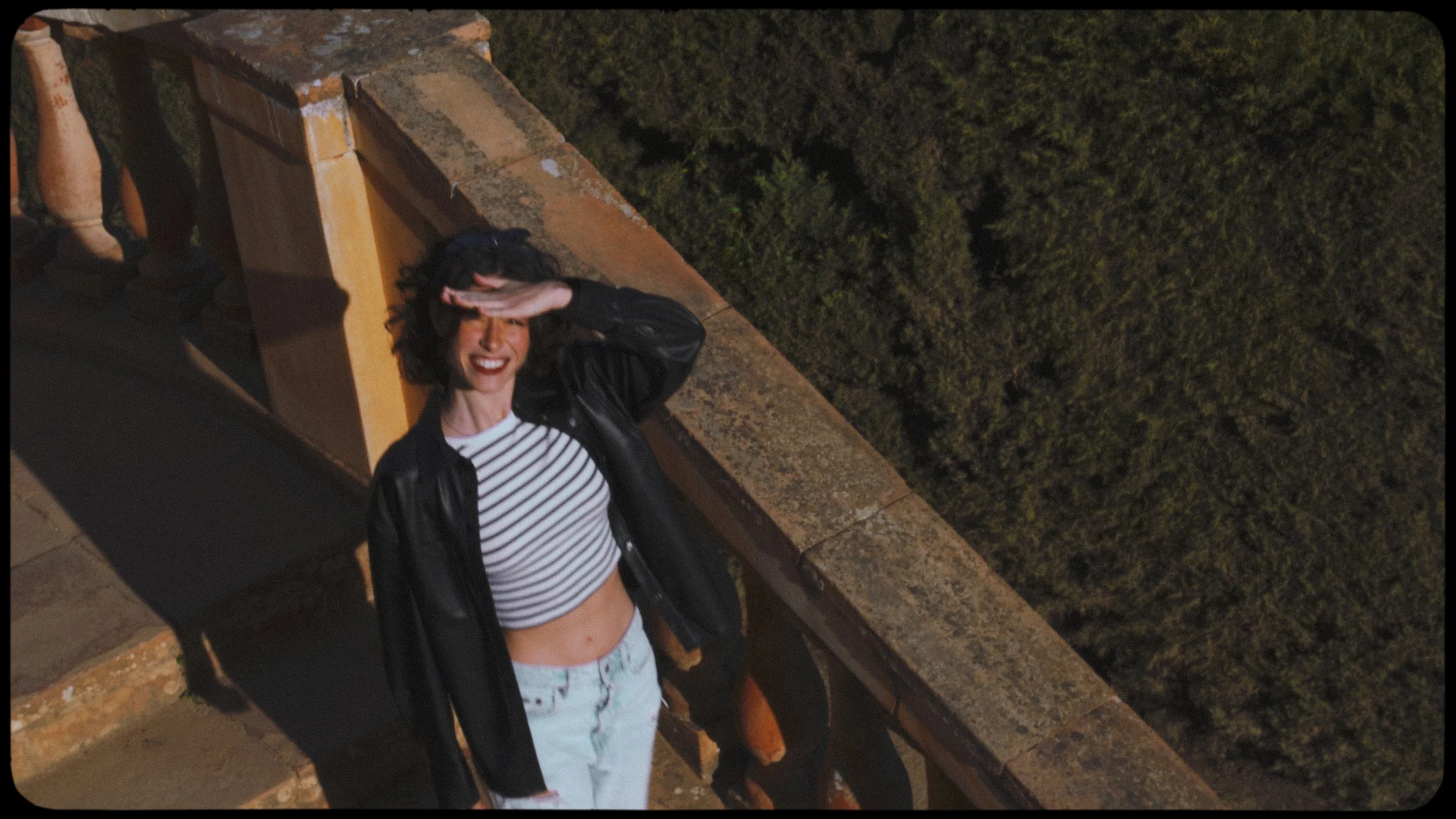 A woman with curly dark hair, wearing a black leather jacket and a striped crop top, is standing on a balcony, smiling and shielding her eyes from the camera with her hand. Behind her is a stone railing and a background of trees.