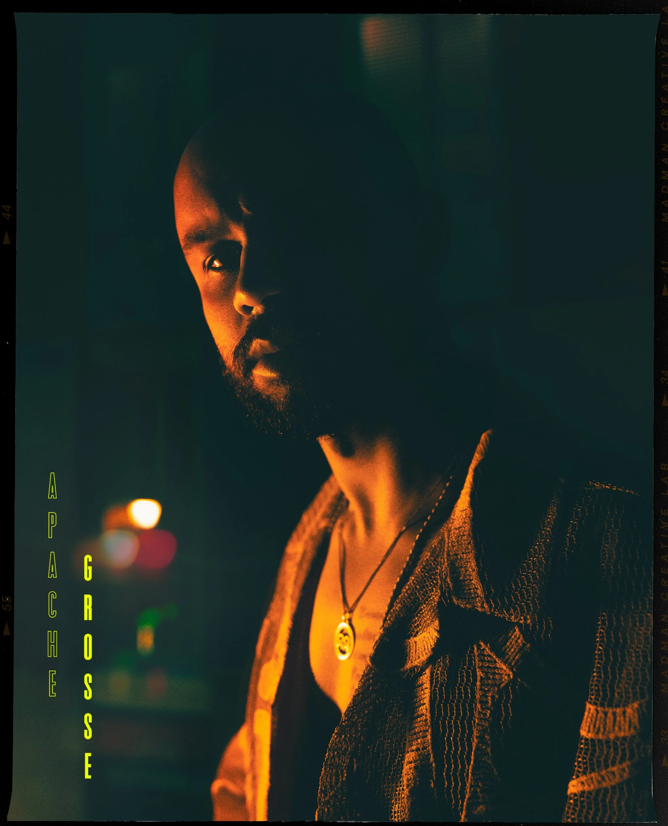 A man with a beard and short hair, lit by warm orange and yellow lighting, wearing a patterned jacket and a necklace with a pendant. The background is dark with colorful blurred lights, giving a moody atmosphere. The word "GROSS" appears vertically i