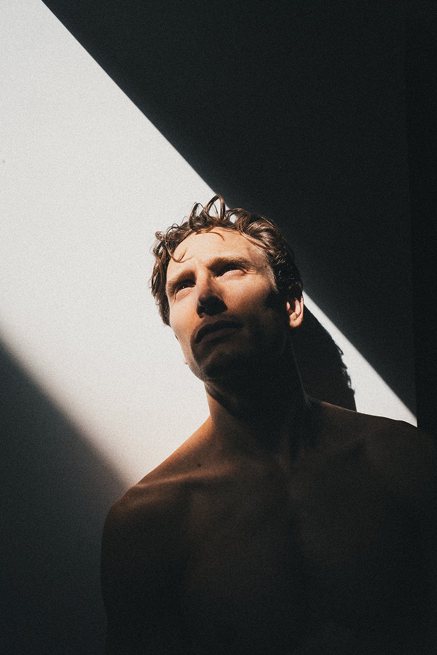 A shirtless man with curly hair looking upwards, illuminated by contrasting light and shadows against a geometric background.