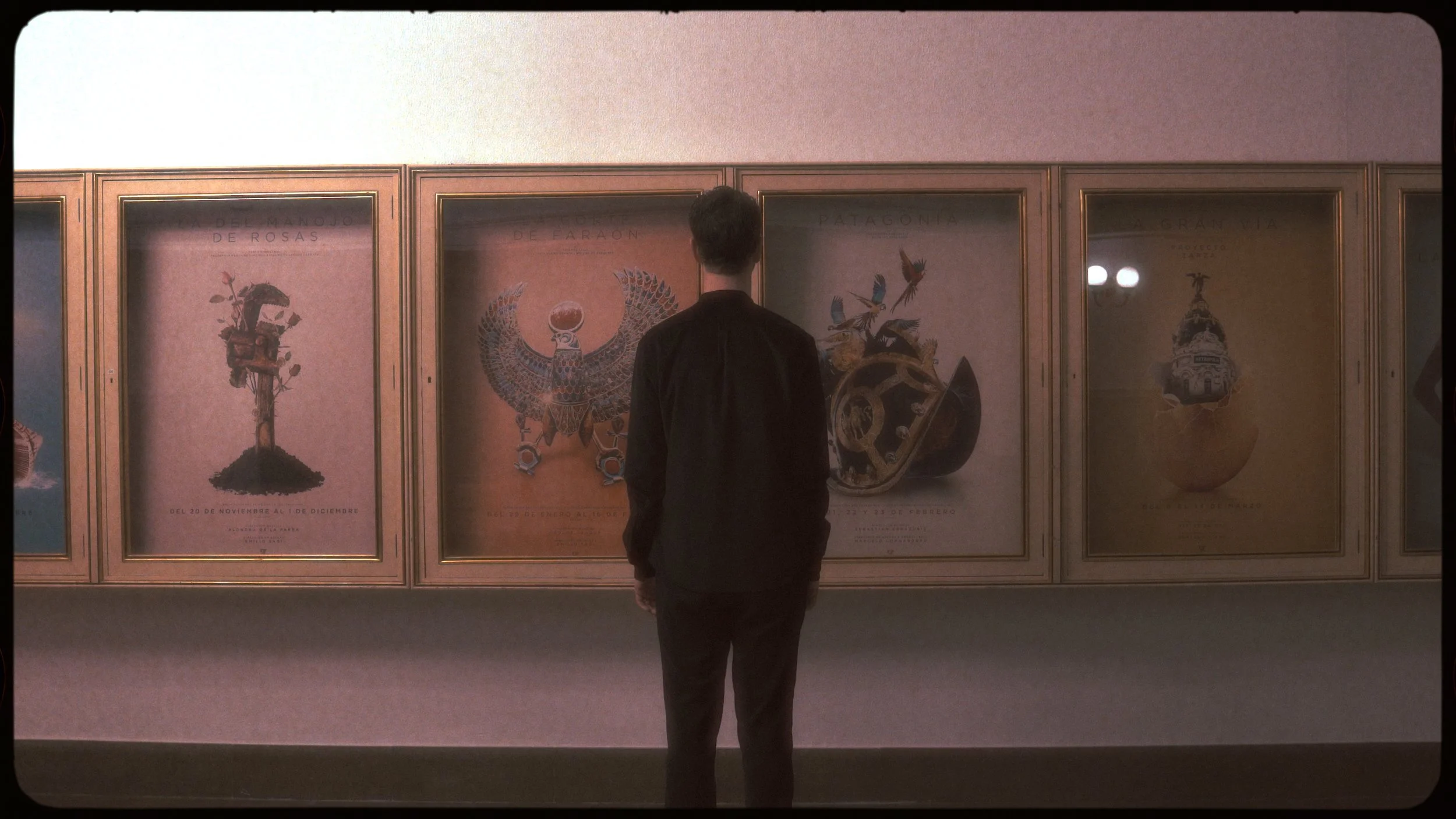 A man stands in front of framed posters of Mexican cultural celebrations or events with text in Spanish, displayed on a gallery wall.
