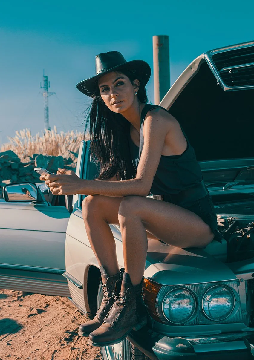 A woman with long dark hair wearing a black hat, a sleeveless top, shorts, and boots sitting on the open hood of a car while holding a cell phone, outdoors with a clear blue sky.