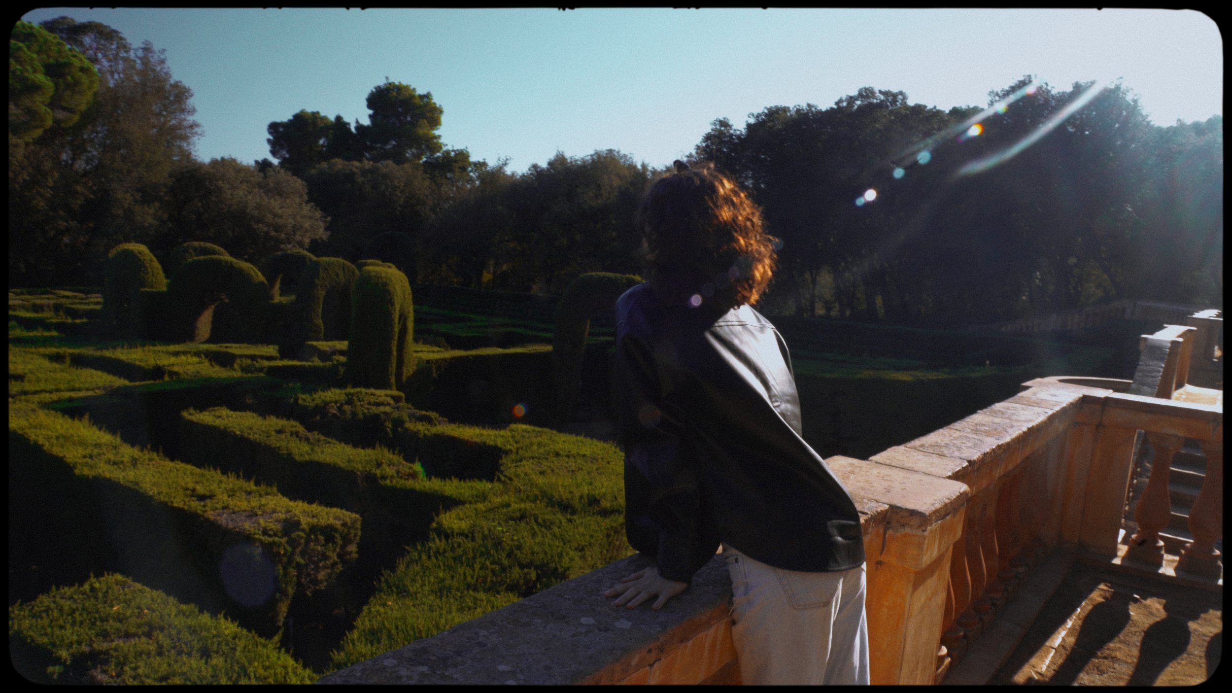 Person leaning on a stone railing overlooking a manicured garden with trees in the background and sunlight creating lens flare.