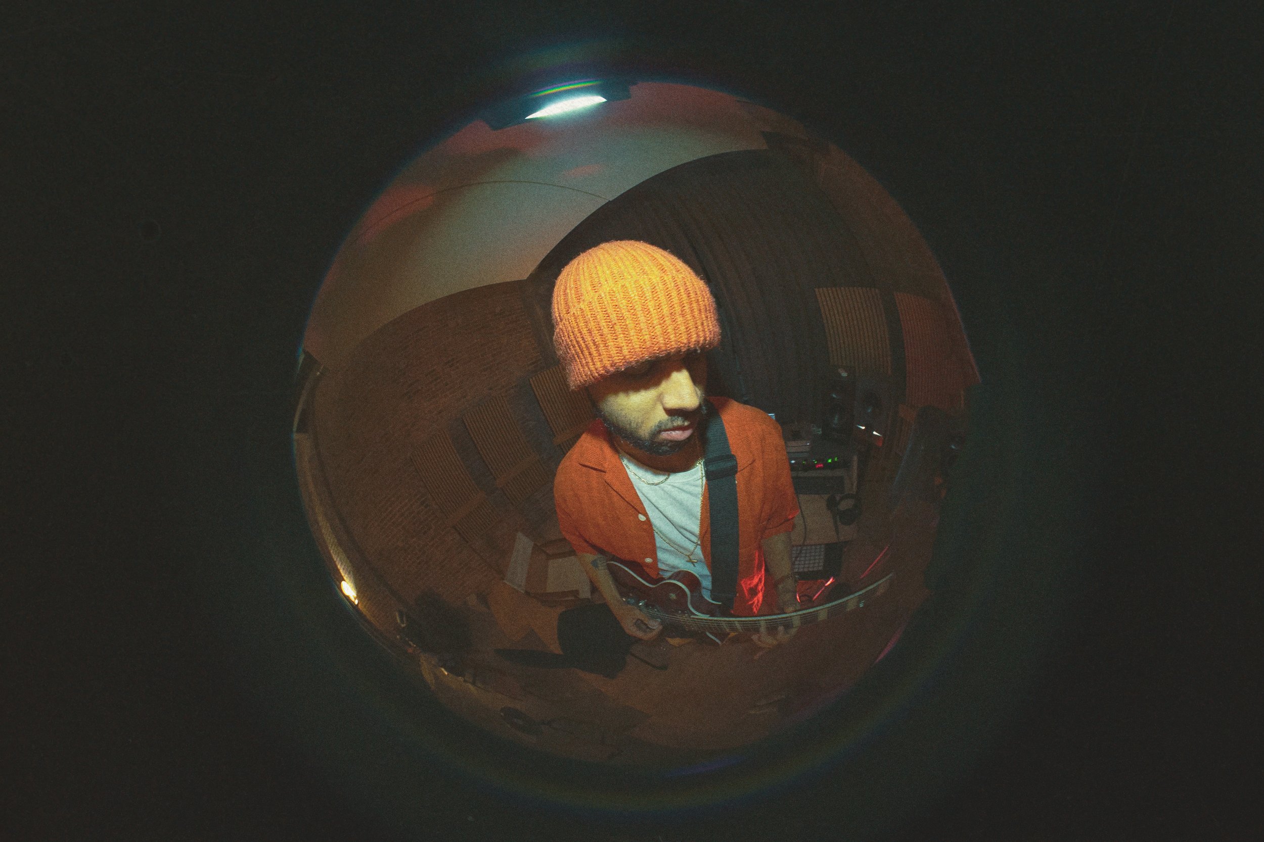 A man wearing an orange shirt and a orange knit cap plays an acoustic guitar, seen through a fisheye lens reflection on a shiny black surface, with studio equipment and acoustic panels in the background.