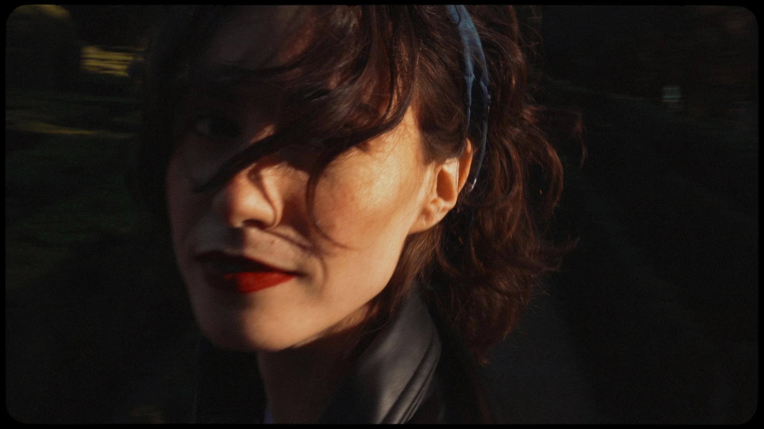 Close-up of a woman with wavy brown hair wearing red lipstick and a dark jacket, with a dark background.