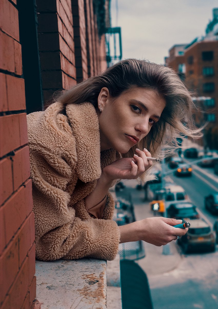 A young woman with blonde hair, wearing a tan fuzzy coat, leaning on a brick wall, holding a cigarette in one hand and a lighter in the other, with a city street in the background.